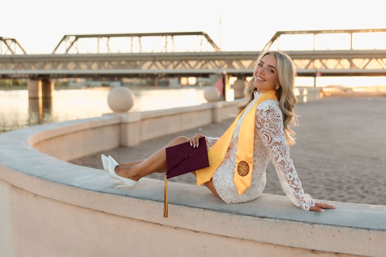 Smiling blonde graduate in lace dress with gold ASU stole sitting outdoors holding maroon diploma cover.
