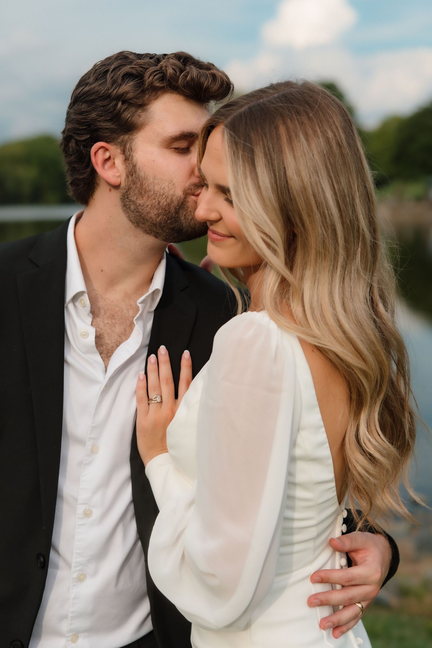 Groom tenderly kissing bride on temple near lakeside, bride in white dress displaying engagement ring on chest.