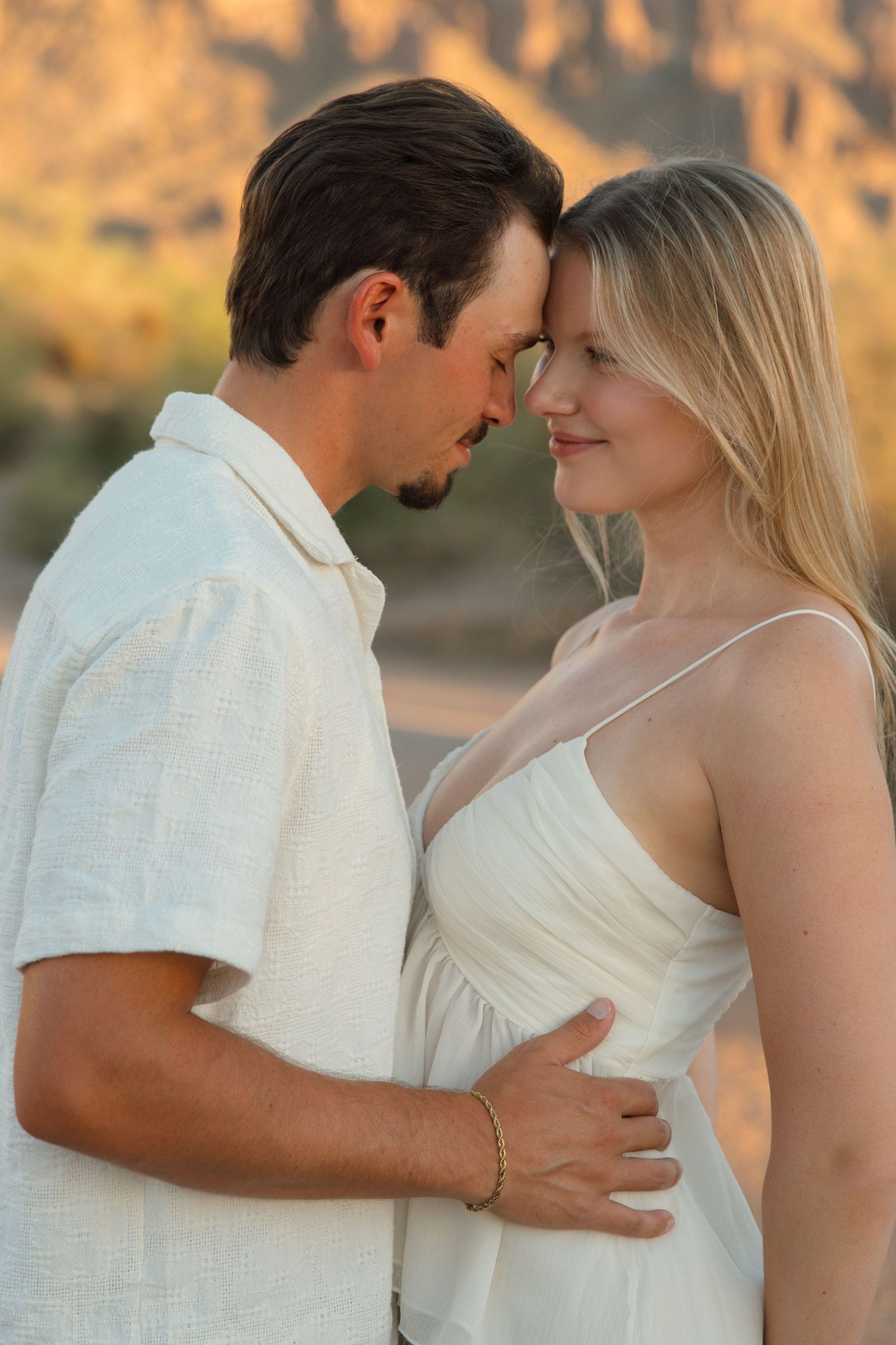 Romantic couple in white outfits touching foreheads tenderly outdoors with warm desert mountain backdrop at sunset.