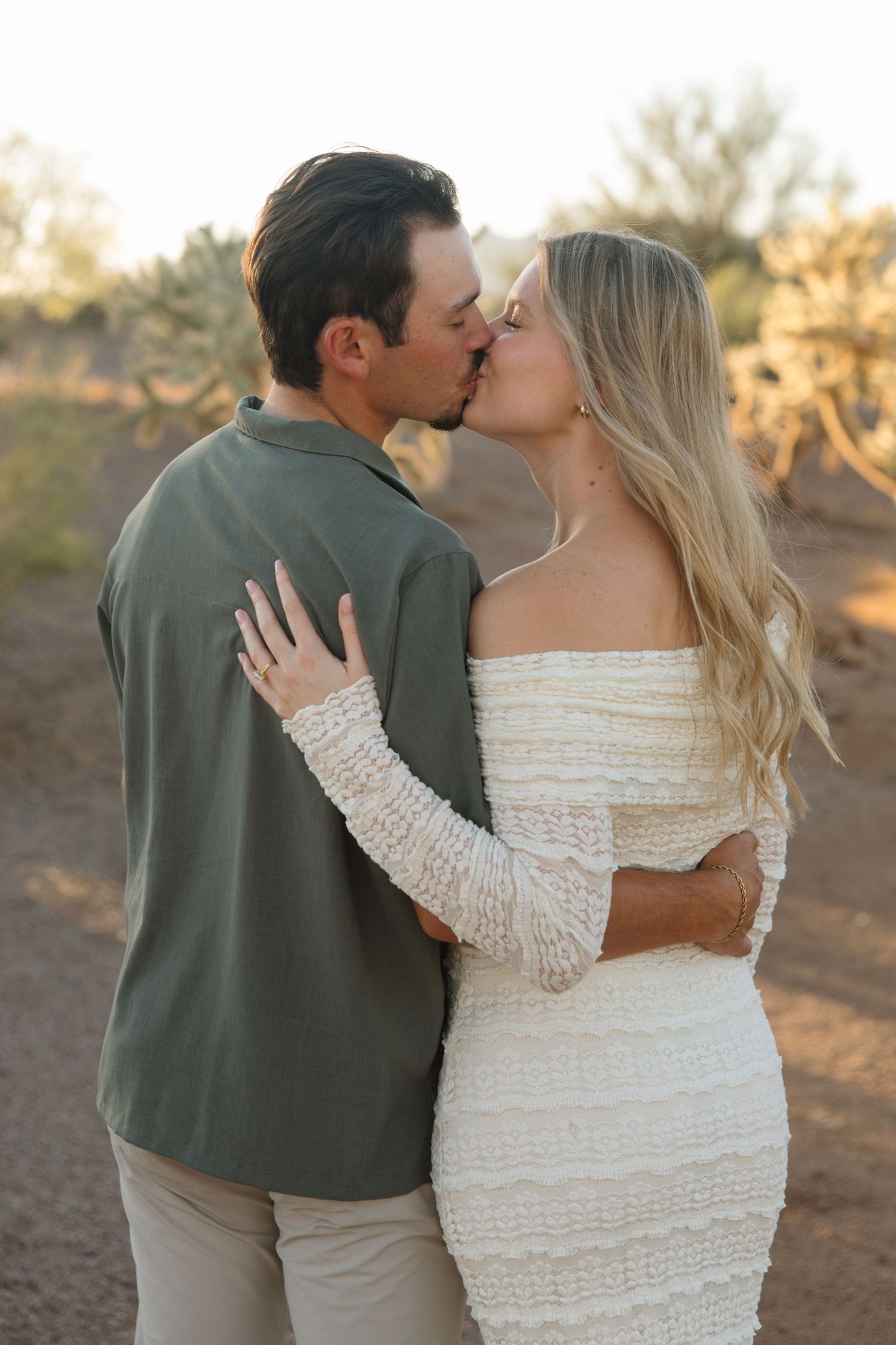 Couple sharing a romantic kiss outdoors in a desert setting, woman in white lace dress, man in olive green shirt.