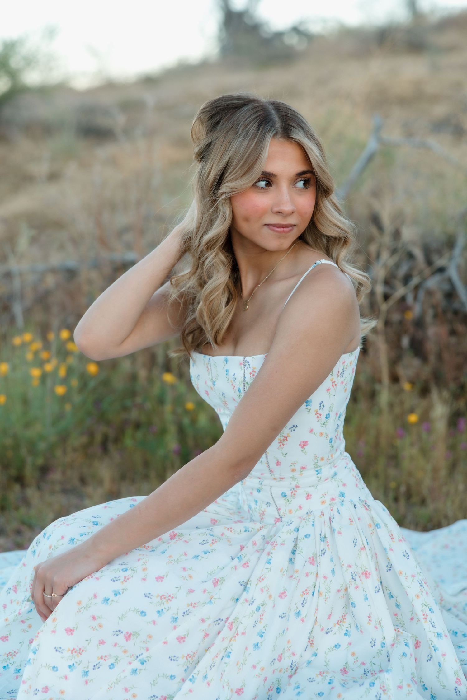 Young woman in floral sundress sitting outdoors in a field, with wavy blonde hair and a gentle smile.