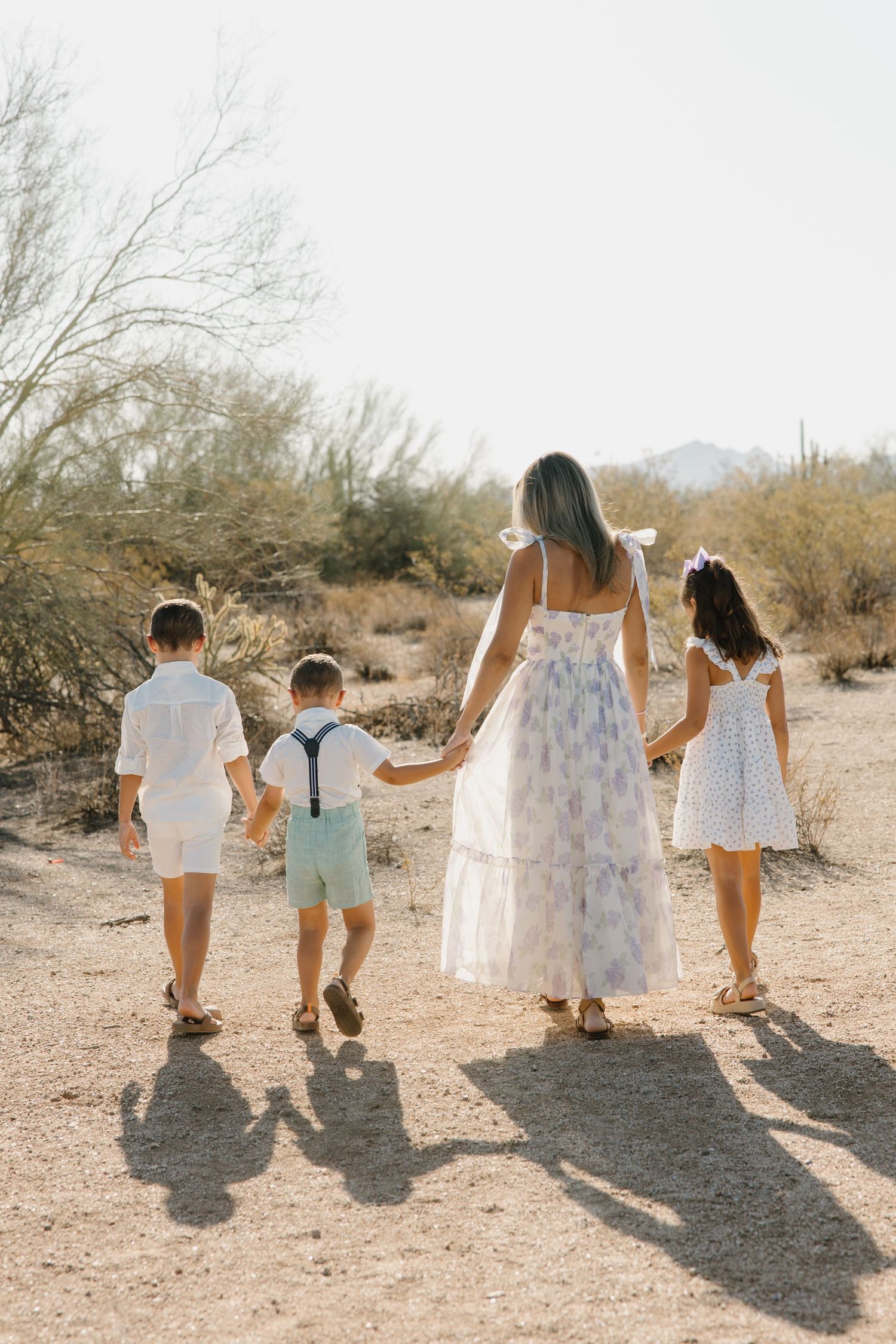 A mother walks hand-in-hand with three children through a desert landscape, all dressed in white and pastel outfits.