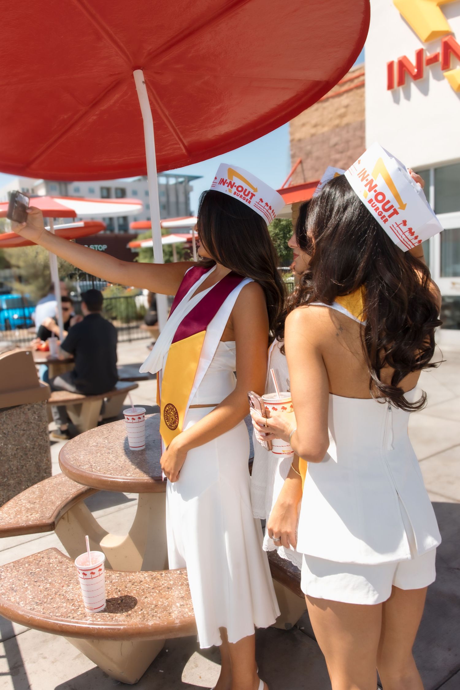 Two graduates in white dresses and gold stoles take a selfie at an In-N-Out Burger outdoor seating area.