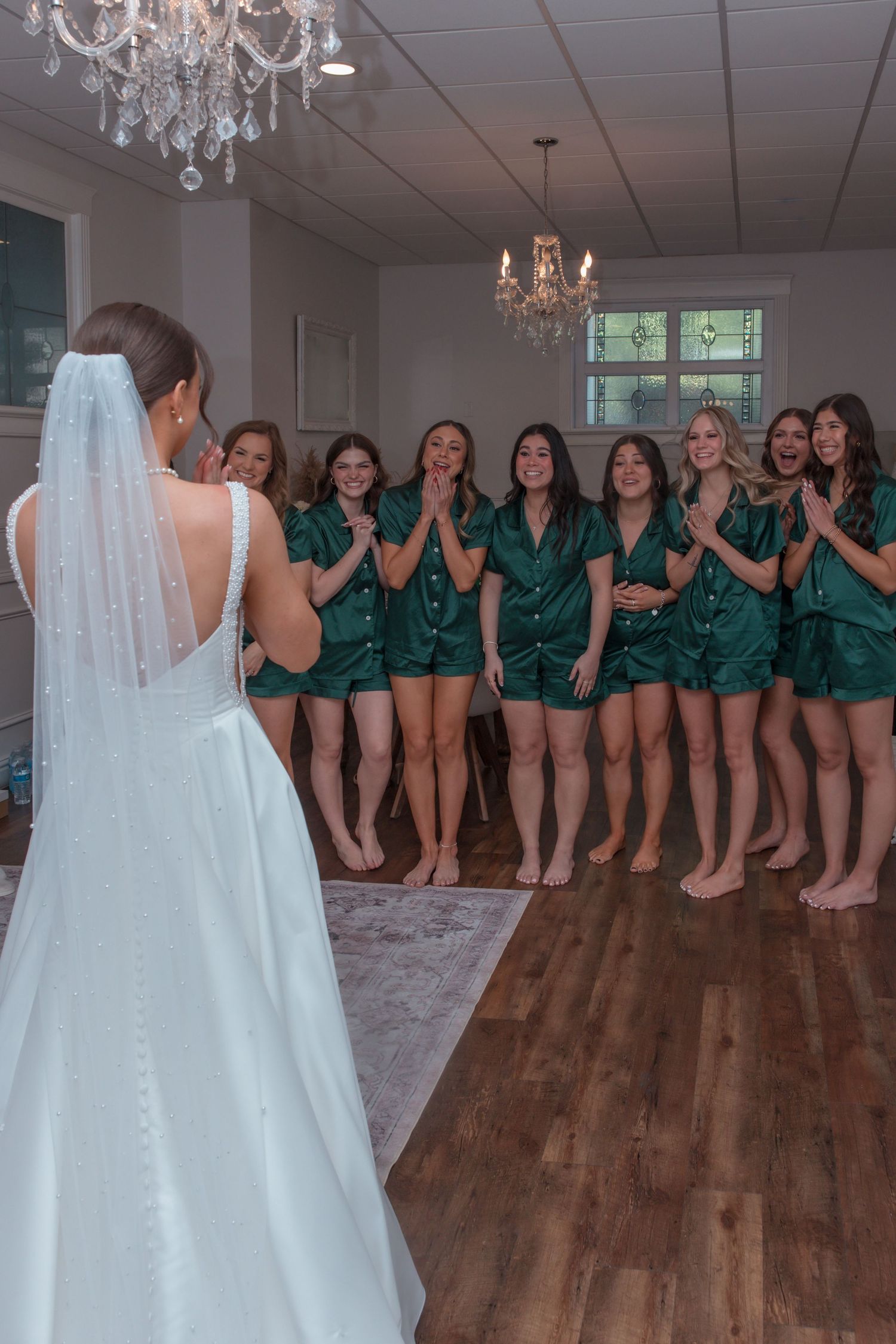 Bridesmaids in green satin pajamas reacting with excitement to bride in white wedding gown and veil.
