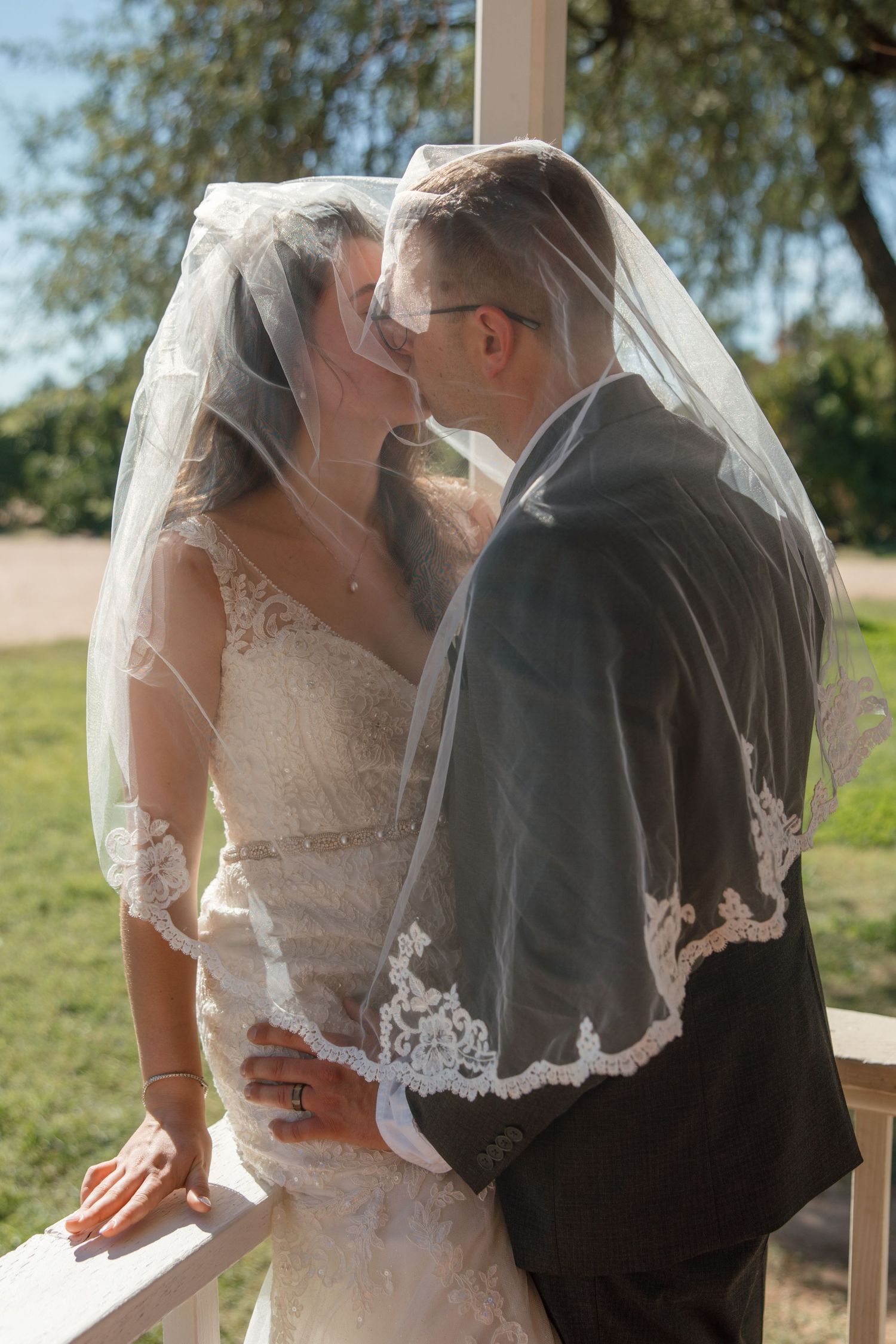 Bride and groom kiss under a flowing lace wedding veil on an outdoor porch on a sunny day.