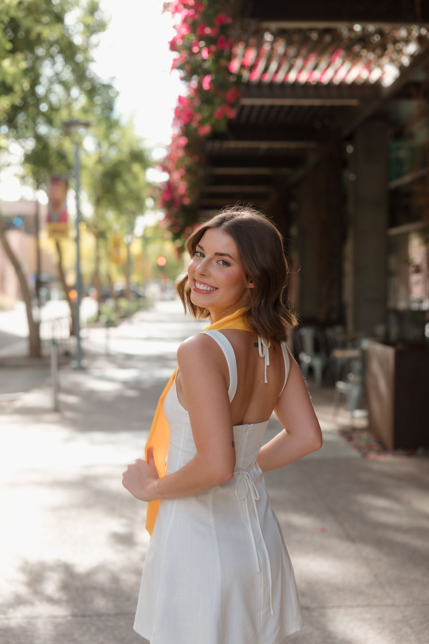 Young woman in white dress with gold sash smiles over shoulder on sunny tree-lined street.
