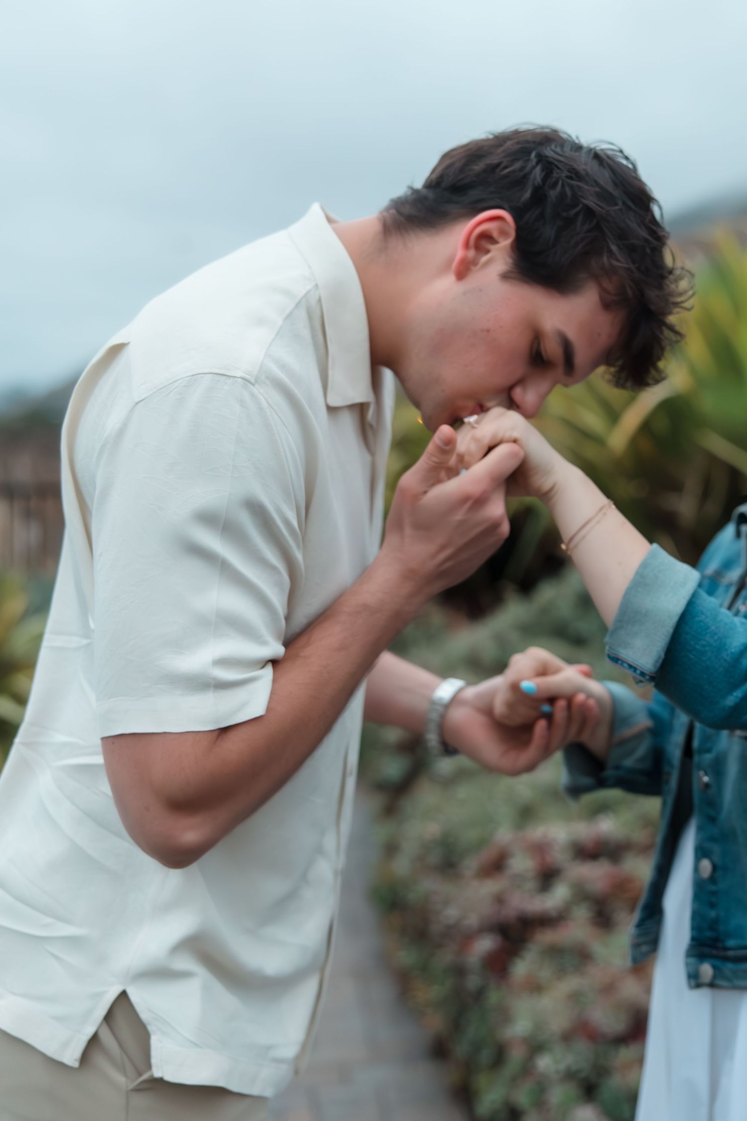 Man tenderly kissing his fiances hand after placing an engagement ring on her finger outdoors.