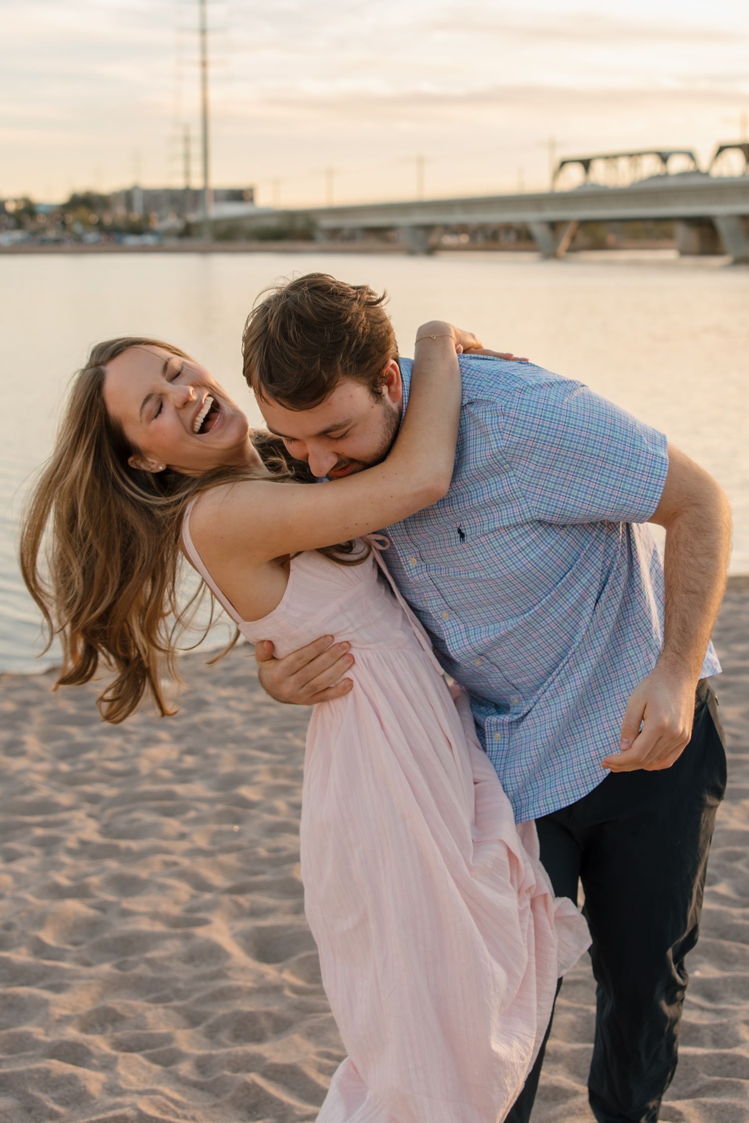 Happy couple embracing and laughing on sandy beach at sunset, woman in pink dress being lifted by man.