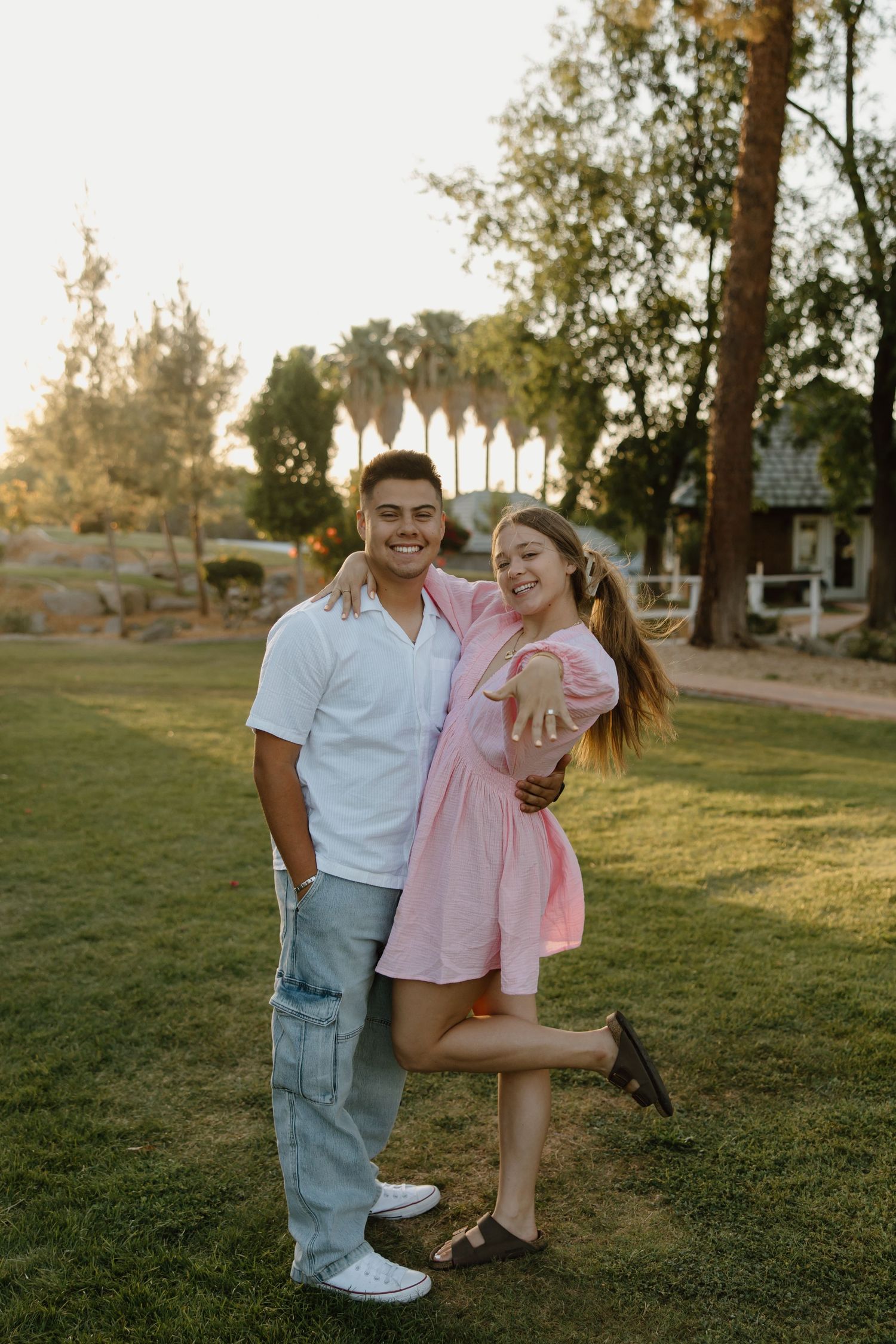 A happy couple poses outdoors at sunset, woman in pink dress showing engagement ring while lifting one leg joyfully.
