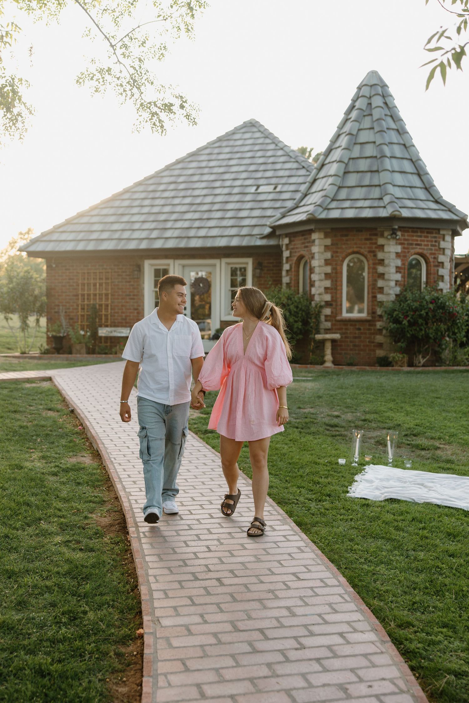 Couple holding hands walking on brick path toward charming brick cottage with pointed roof at sunset.