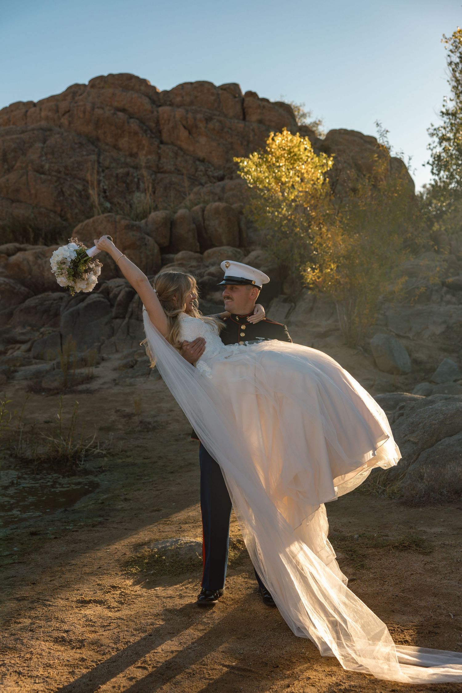 Marine groom lifts bride in flowing white gown as she raises bouquet joyfully in desert setting.