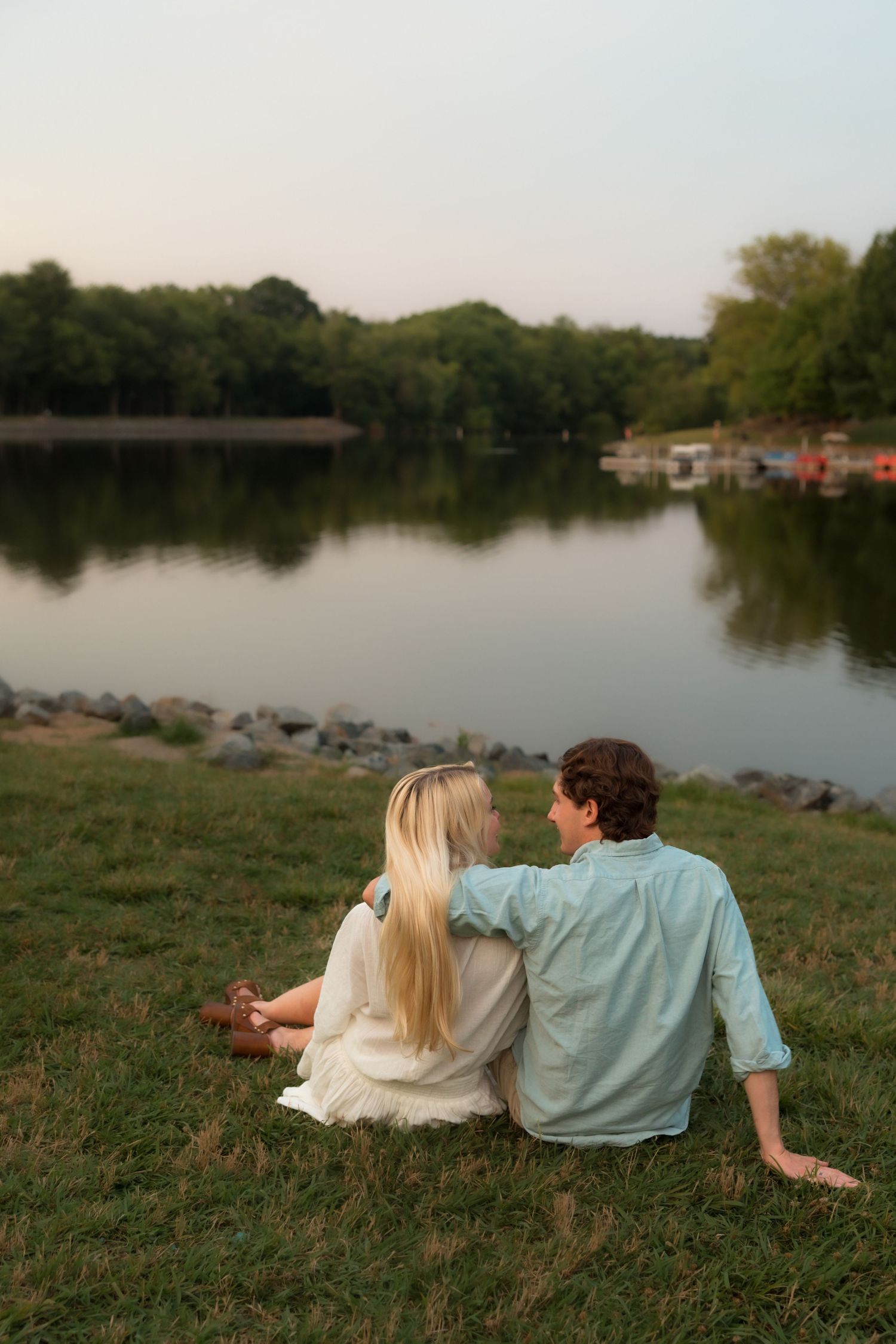 Couple sitting together on grass by a serene lake at sunset, viewed from behind.