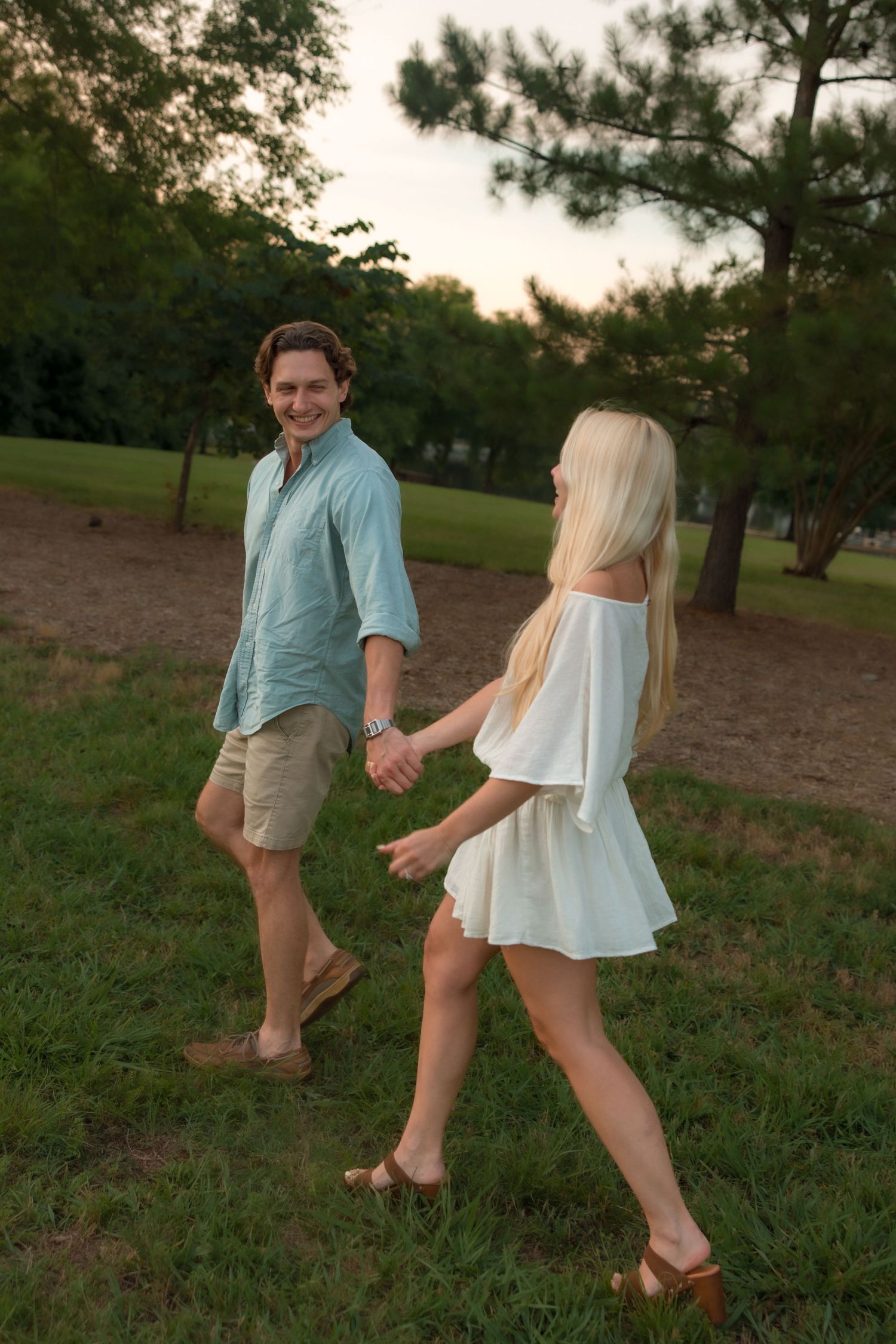 Smiling man leads his blonde partner by the hand across a grassy park at dusk.
