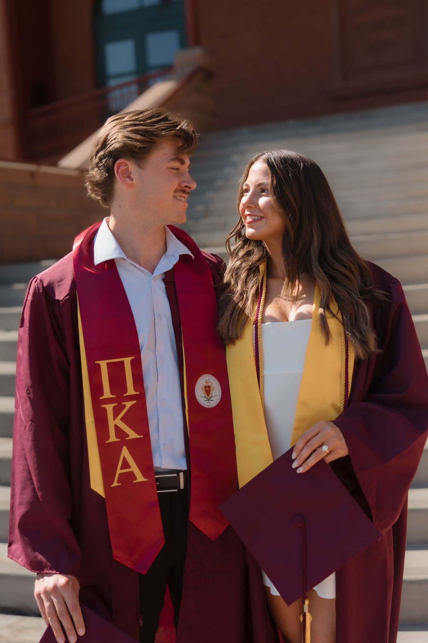 Smiling couple in maroon graduation robes and honor stoles posing on university building steps together.