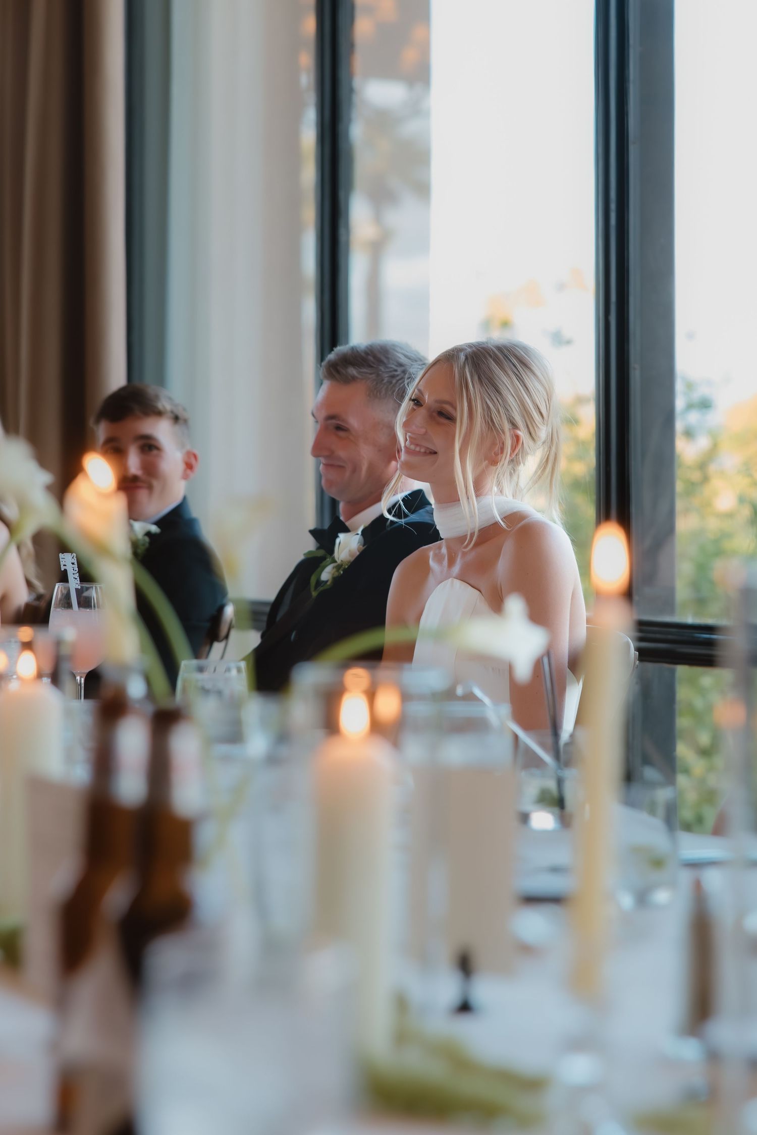 Smiling bride and groom seated at candlelit wedding reception head table with floral arrangements.