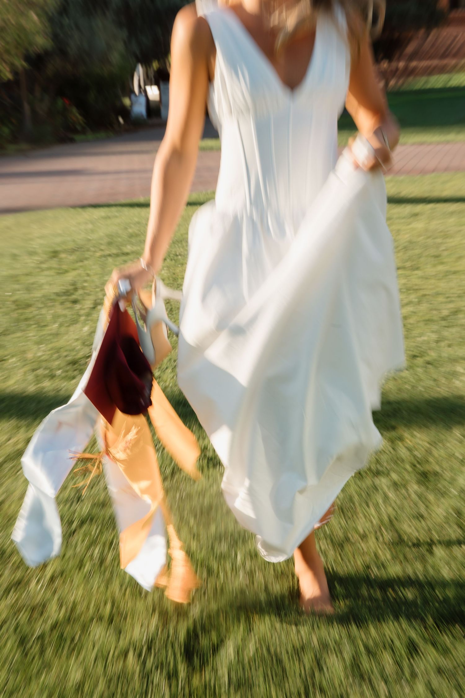 Woman in flowing white dress walking on grass holding maroon graduation cap and gold stole.