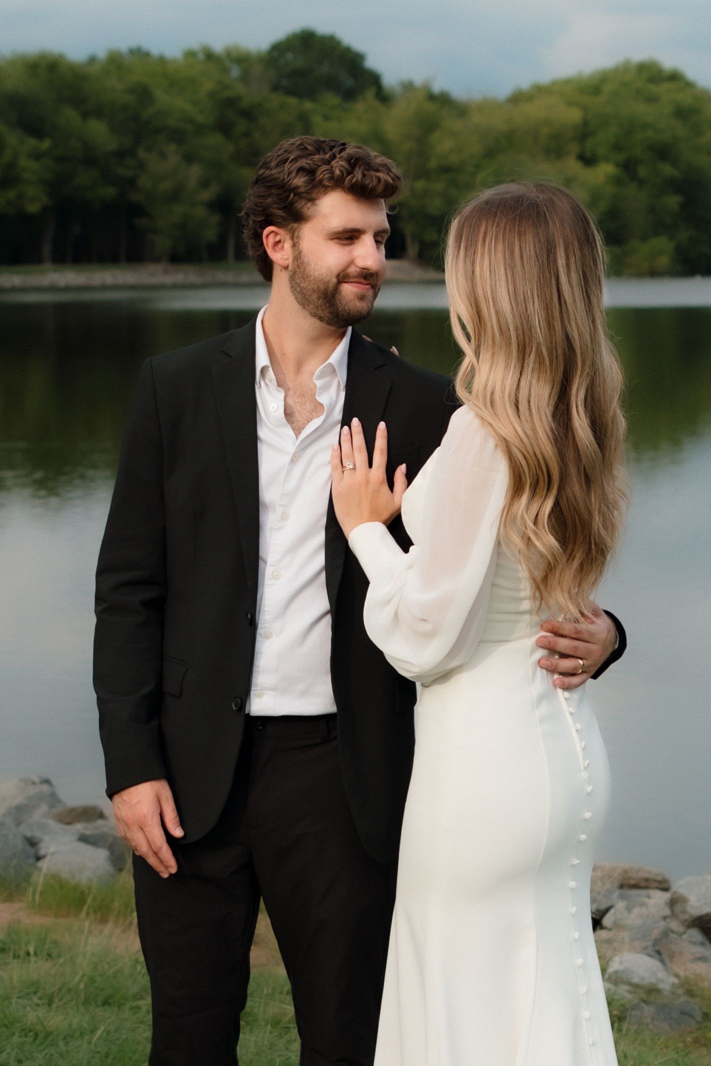 A bride in a white button-back gown shows her ring while embracing a smiling groom in a black suit by a lake.