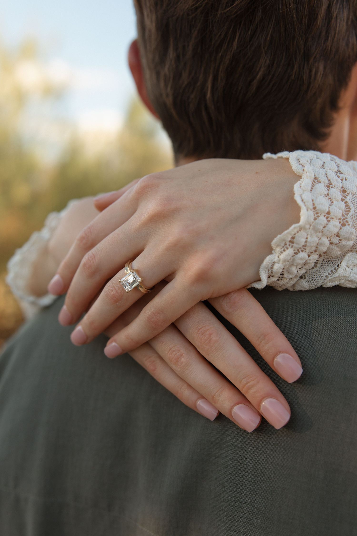 Woman's hand with elegant diamond engagement ring resting on man's shoulder, wearing lace sleeve.