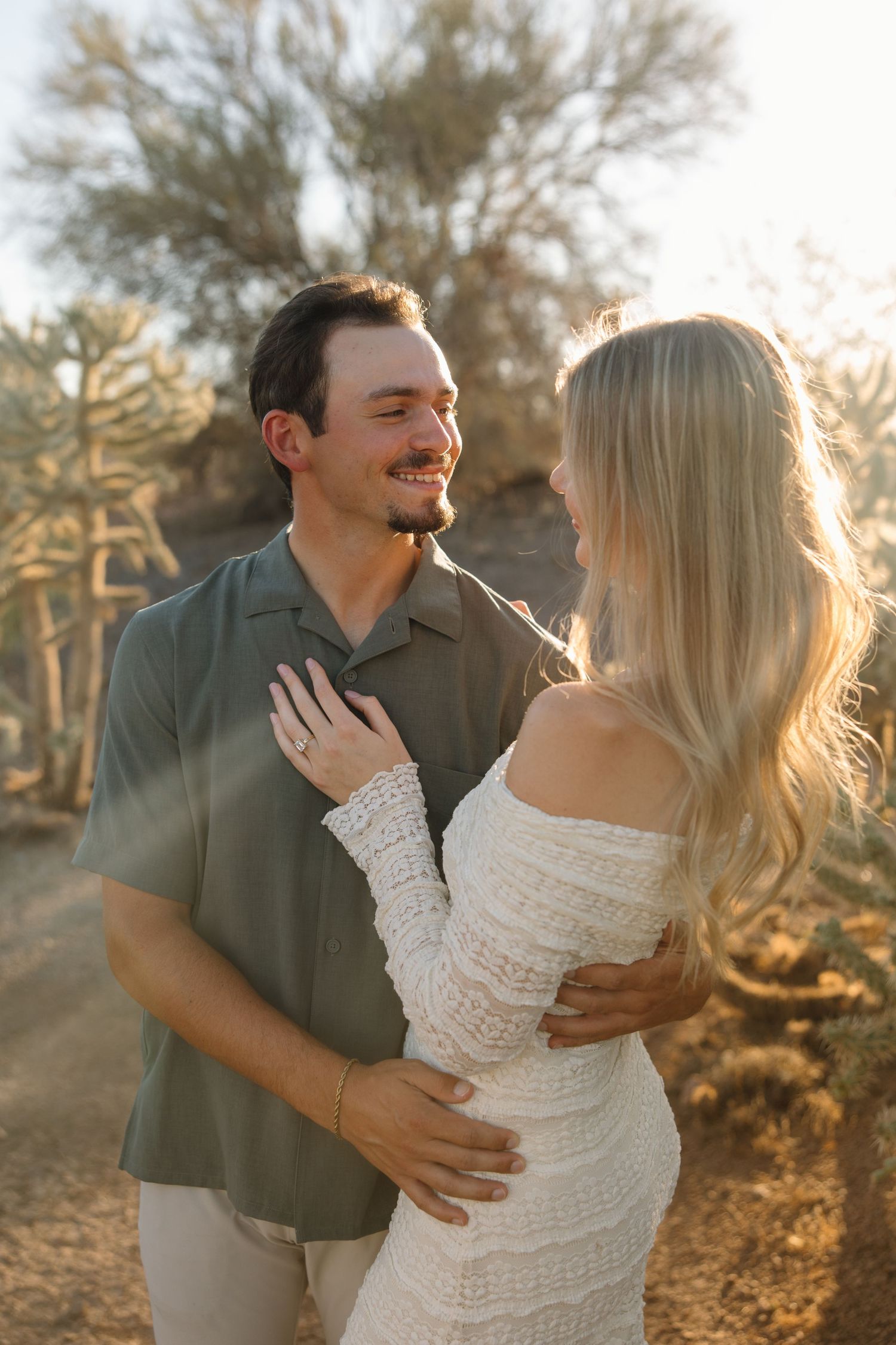 Couple embracing and smiling at each other during golden hour in a desert landscape, woman in white lace dress.