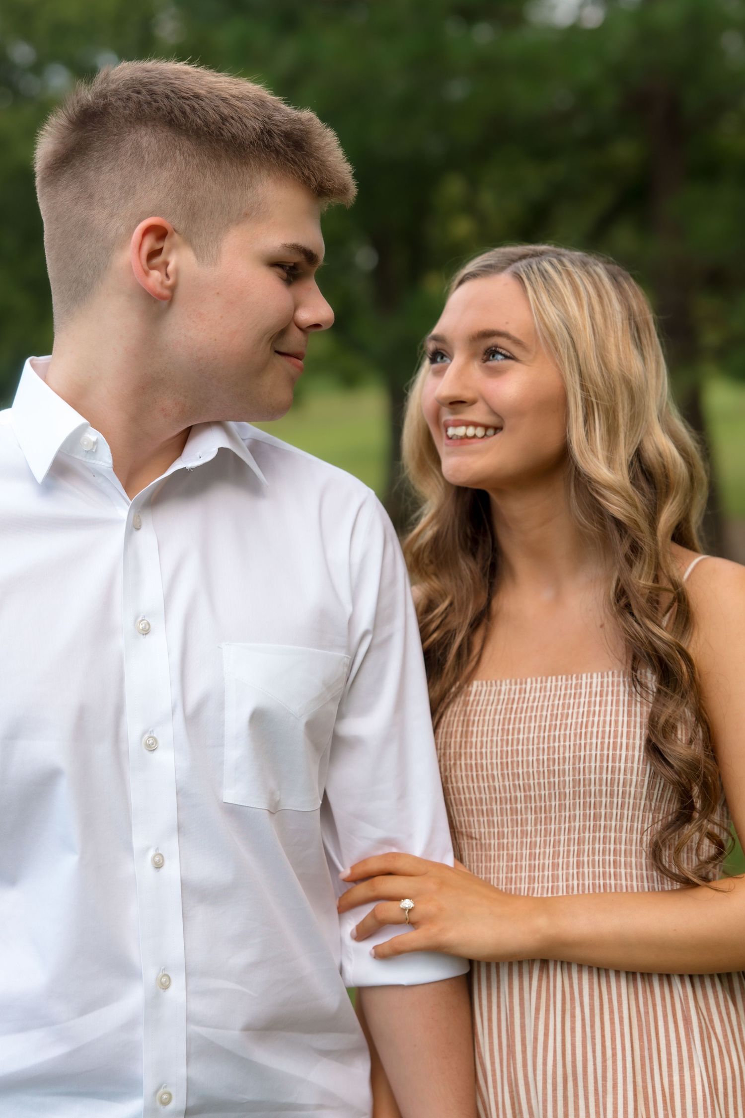 Young couple smiling at each other outdoors, woman in tan dress and man in white shirt, green trees background.