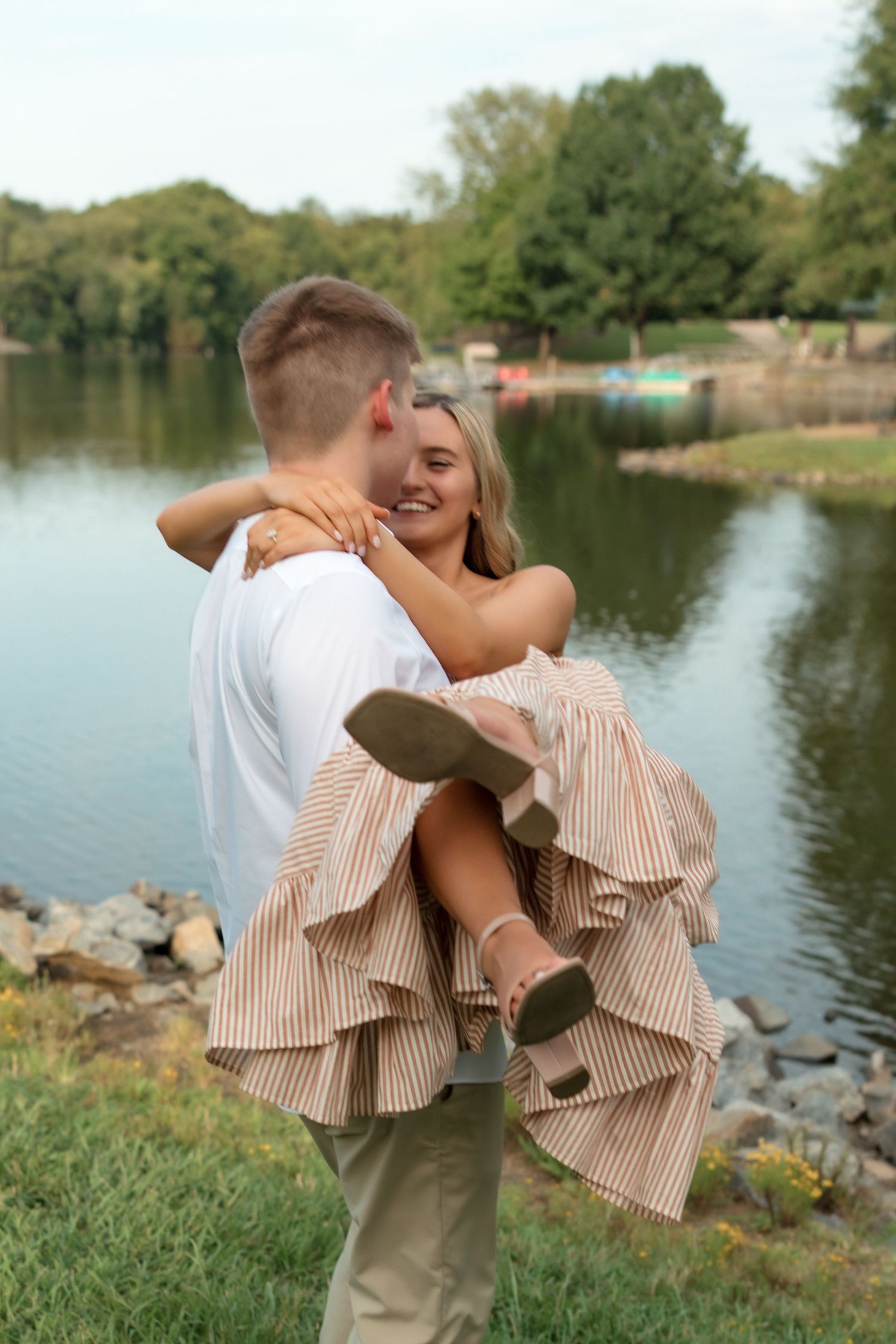 A man lifts a smiling blonde woman in a striped dress by a serene lake surrounded by lush green trees.