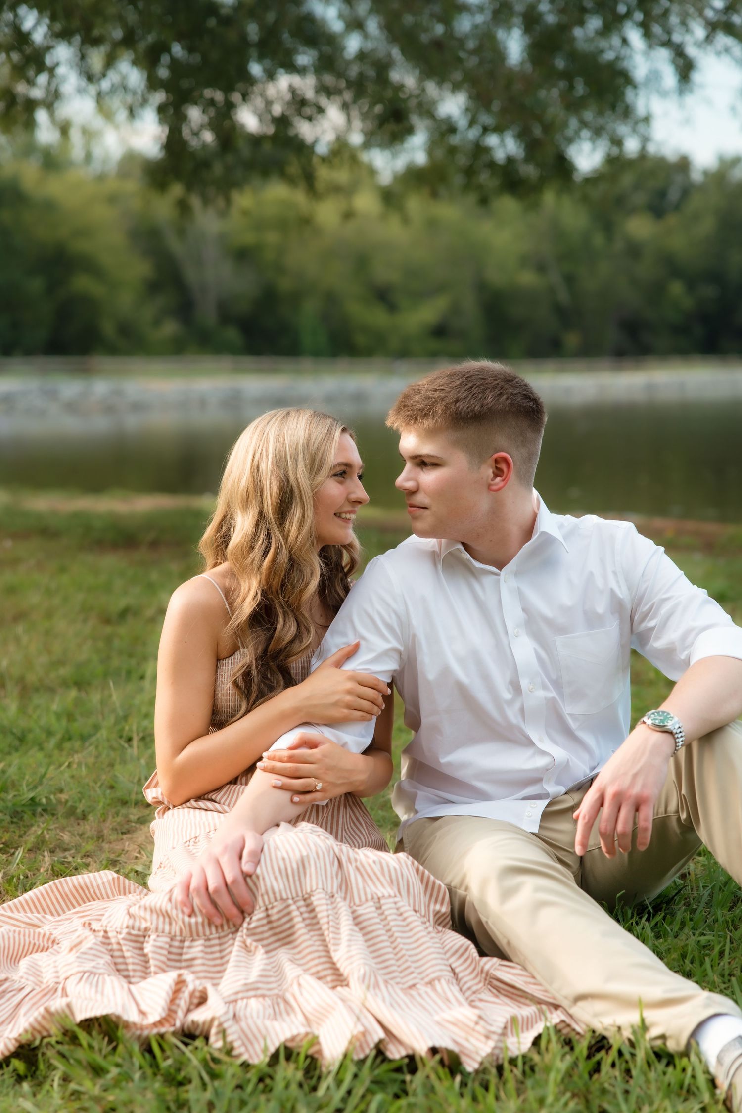 Young couple sitting on a blanket by a lake, smiling at each other in a lush green park setting.