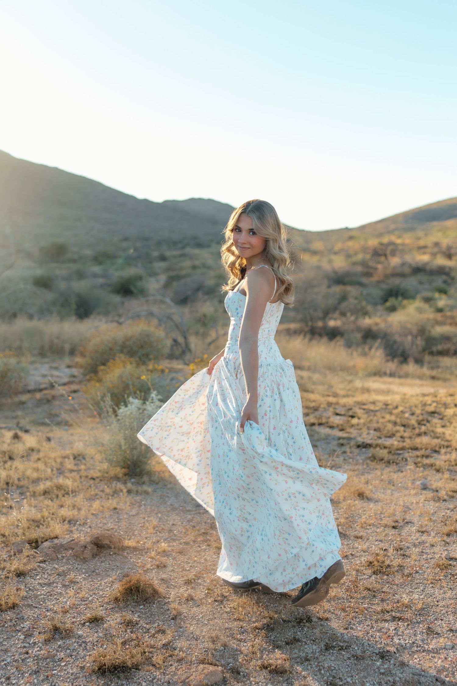 A blonde woman in a flowing white lace dress twirls in a sunlit desert landscape with golden hills.