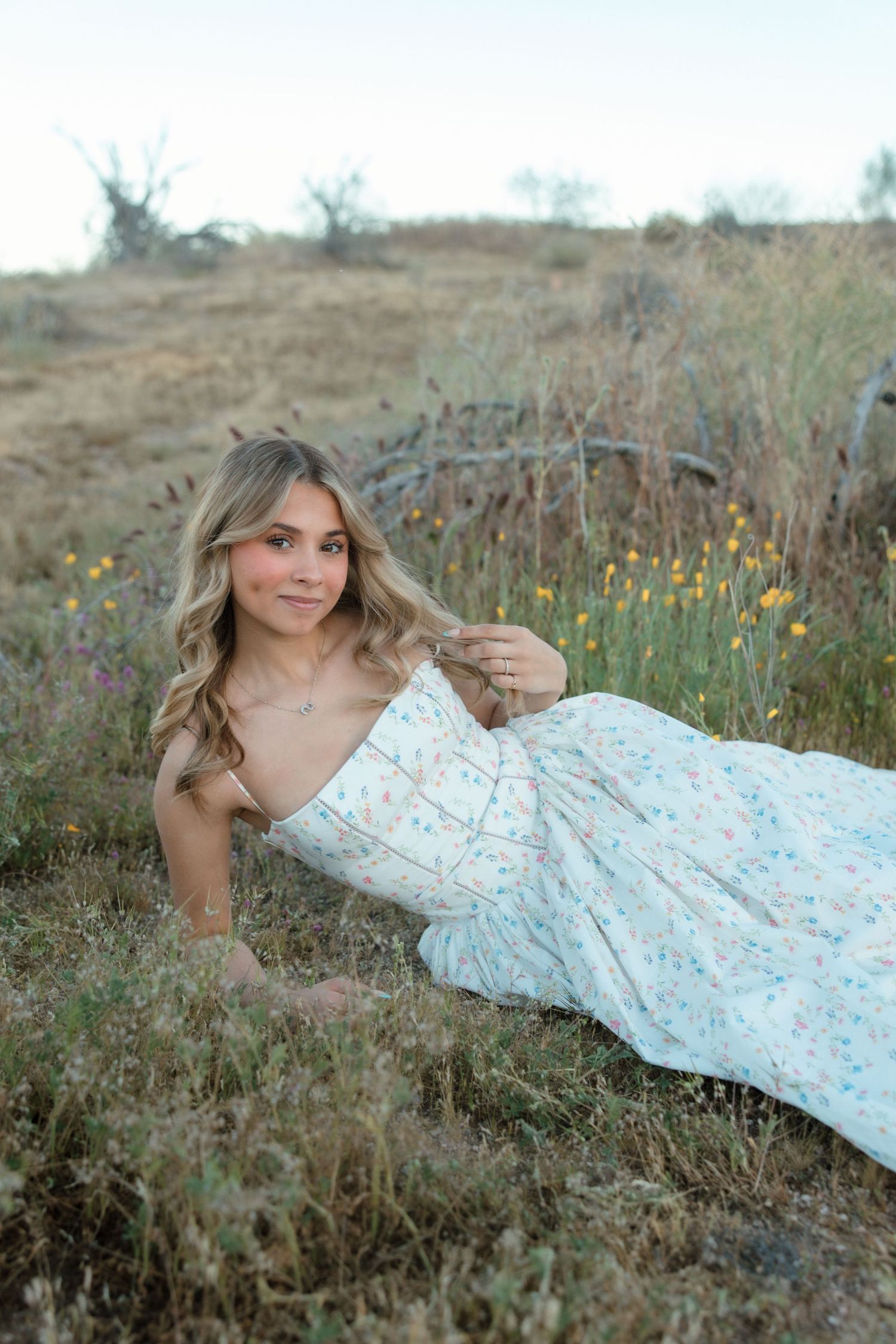 Young woman in floral dress lying in a golden meadow with wildflowers, smiling softly at camera.