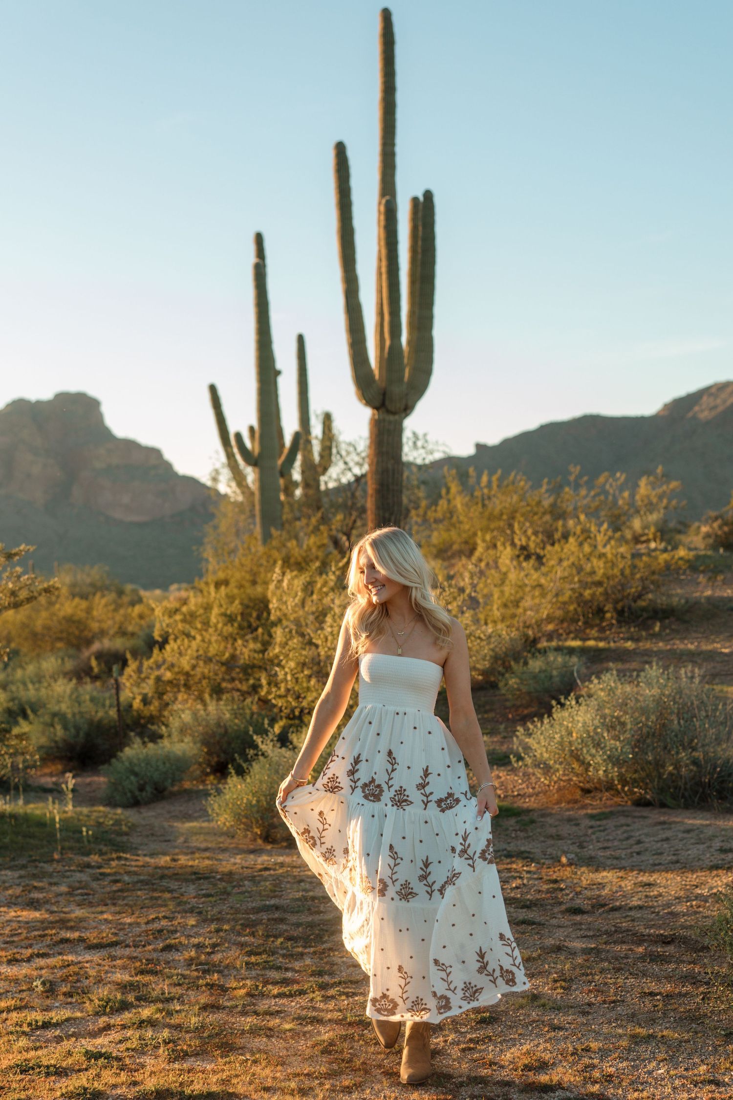 Blonde woman in white embroidered maxi dress walking in Arizona desert with saguaro cacti and mountains at golden hour.