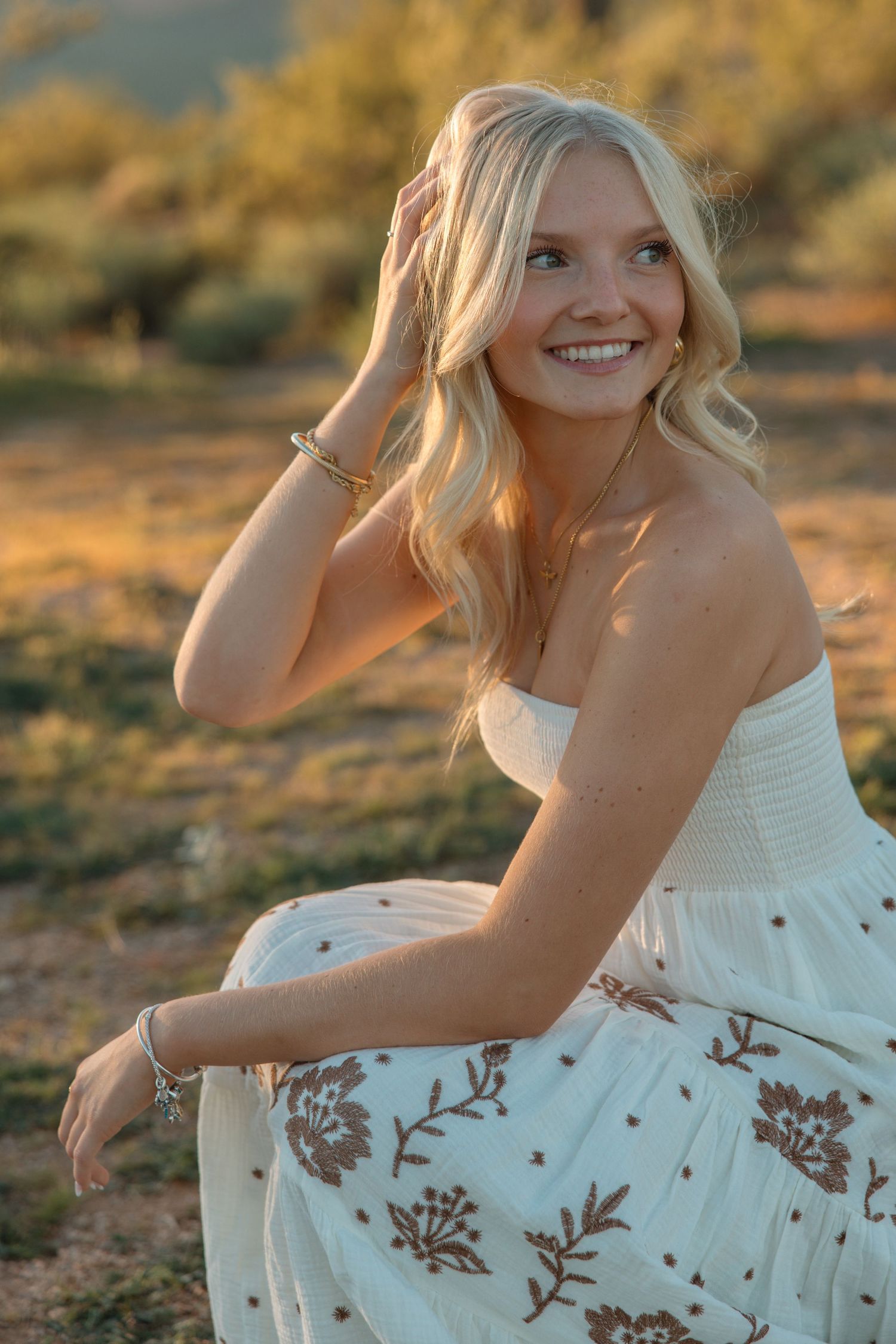 Smiling blonde woman in white floral strapless dress sitting outdoors in golden sunset light.