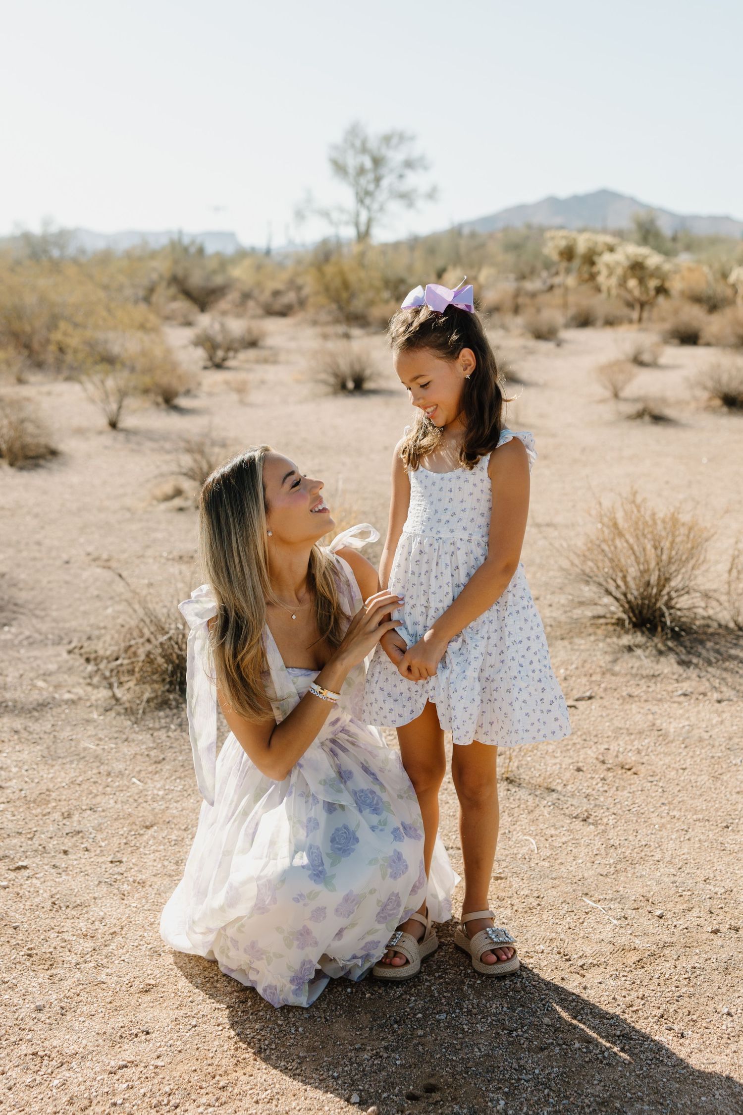 A mother kneels in a desert landscape smiling at her young daughter in matching floral dresses.