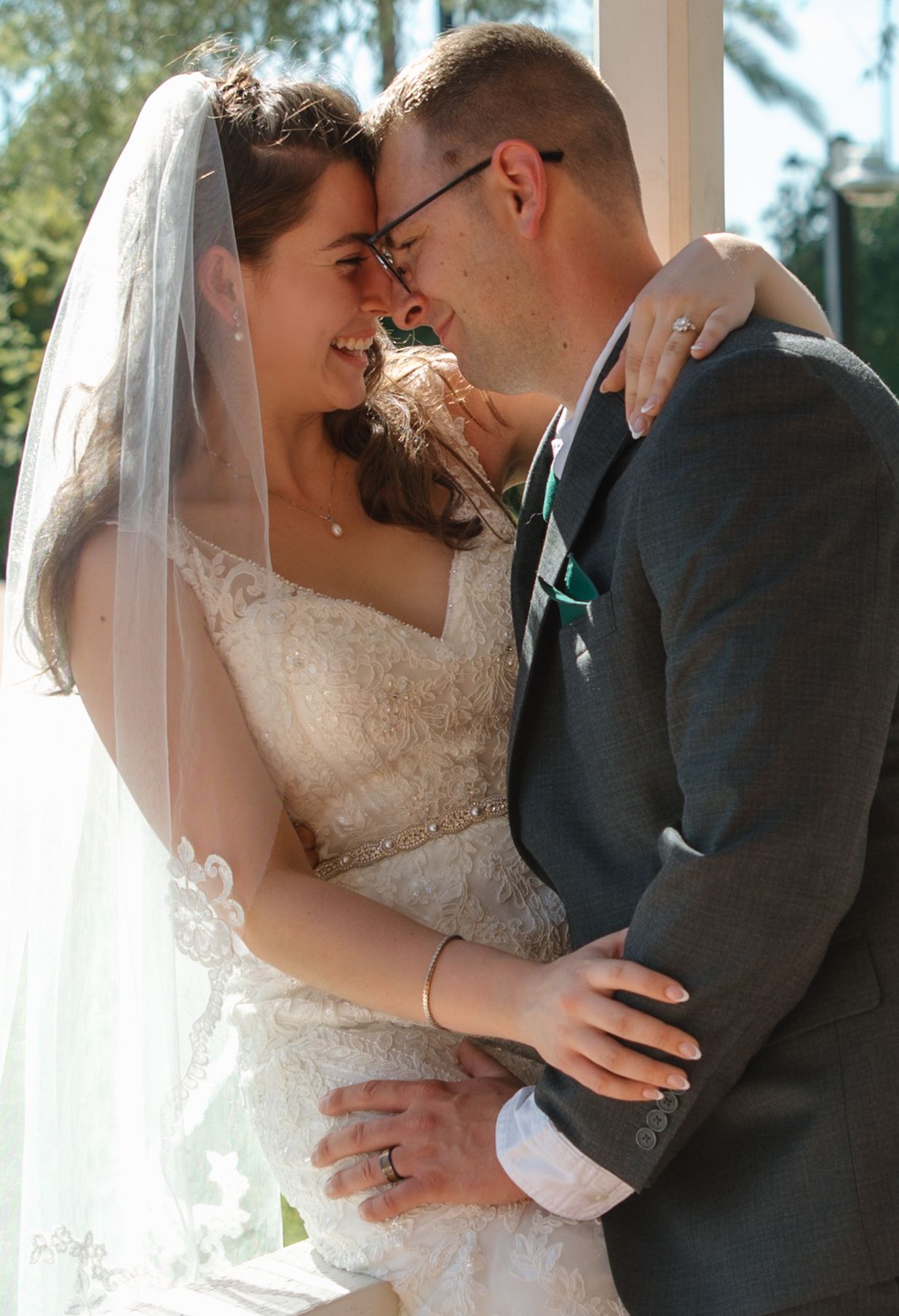 A bride and groom share a romantic moment, smiling at each other, she in a lace gown with veil, he in a gray suit.