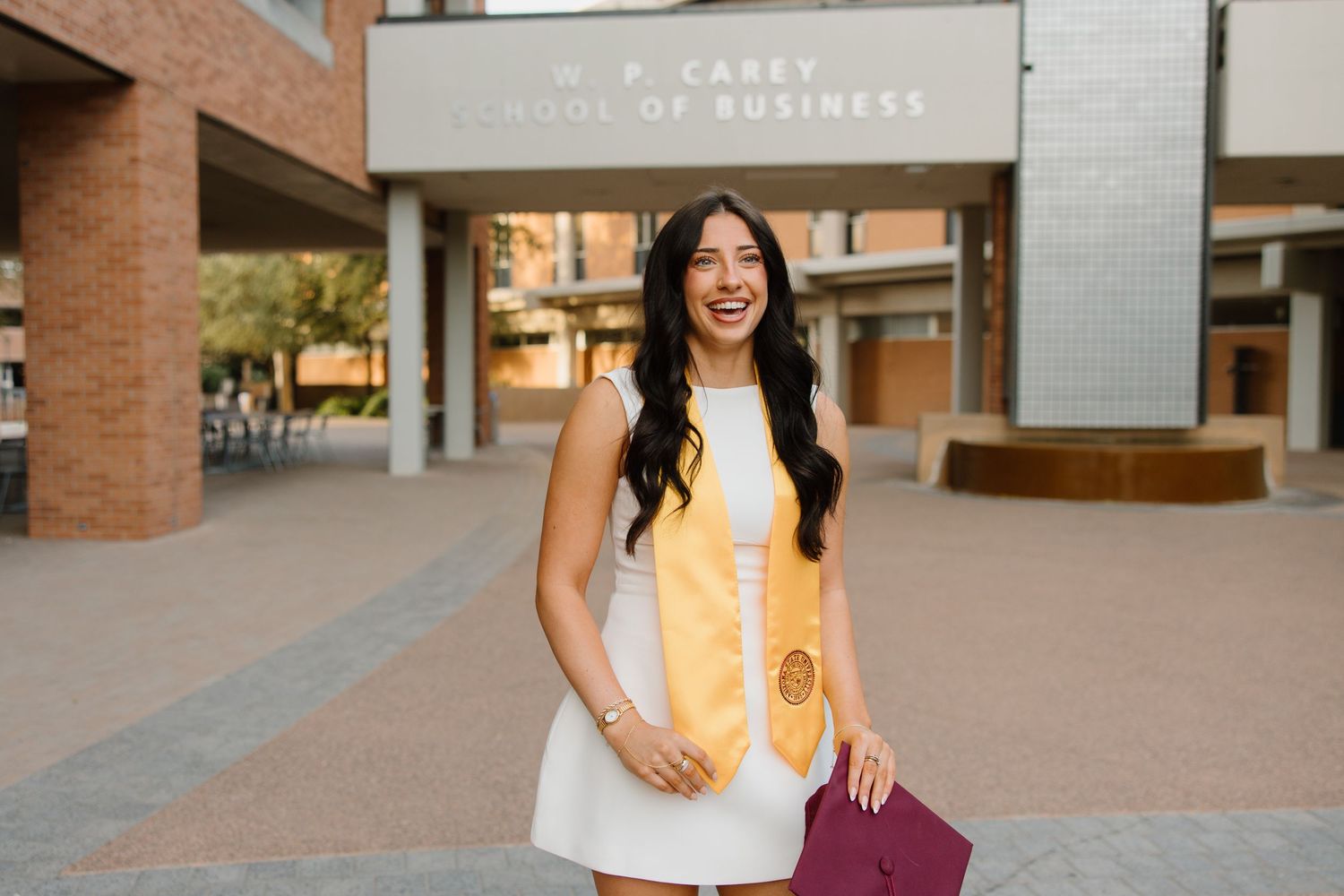 Smiling graduate in white dress and gold stole holds diploma outside W.P. Carey School of Business.