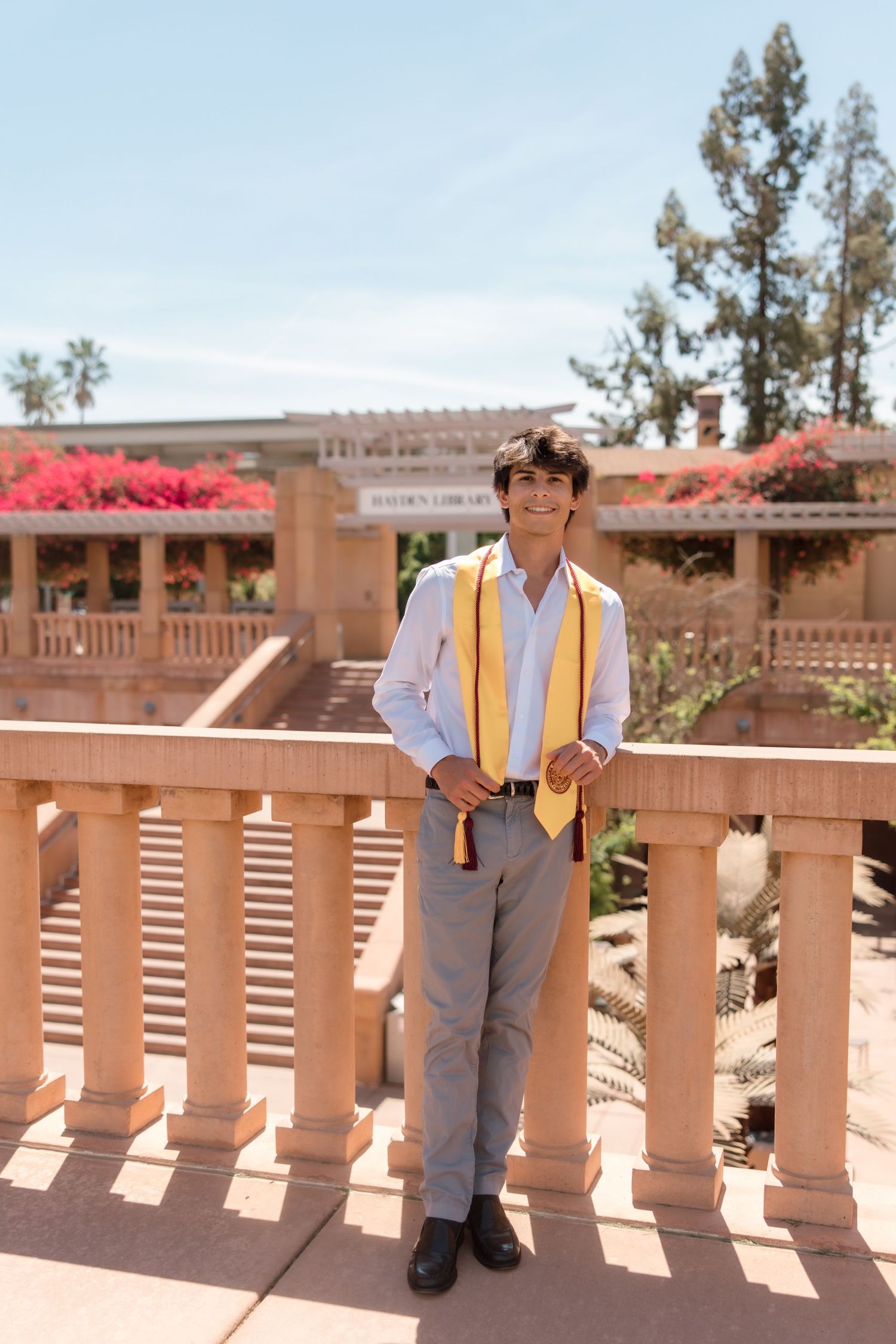 Young man in white shirt and gold graduation stole smiling on a terrace with pink flowers and trees.