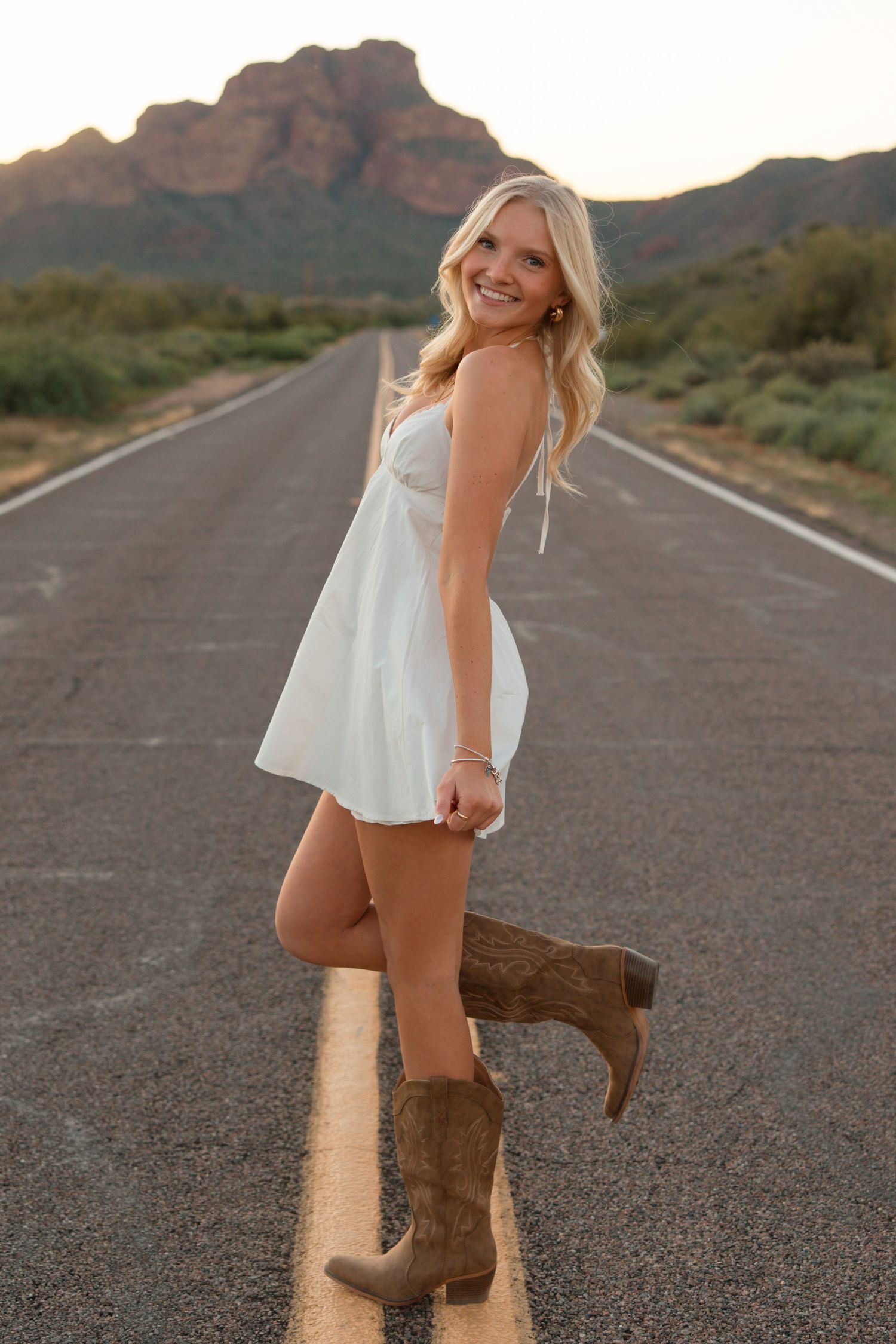 Blonde woman in white dress and cowboy boots posing on open road with mountain backdrop.