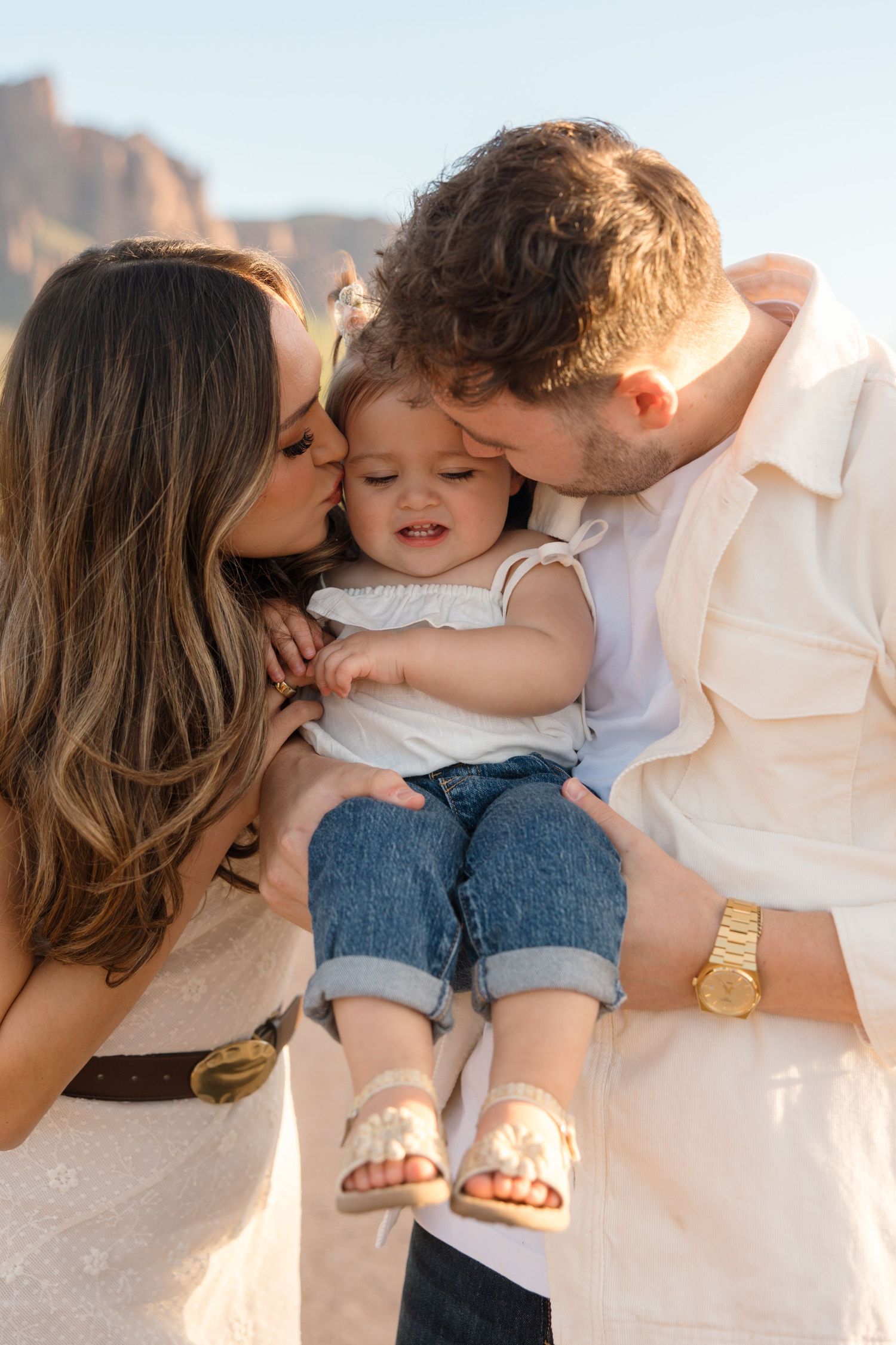 Parents kissing their baby girl on the cheeks while holding her in a desert landscape.