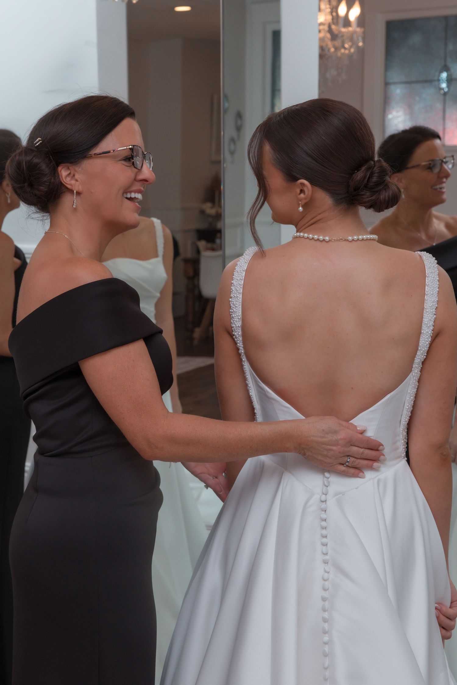 Woman in black dress smiling at bride in backless pearl-button wedding gown during bridal preparations.