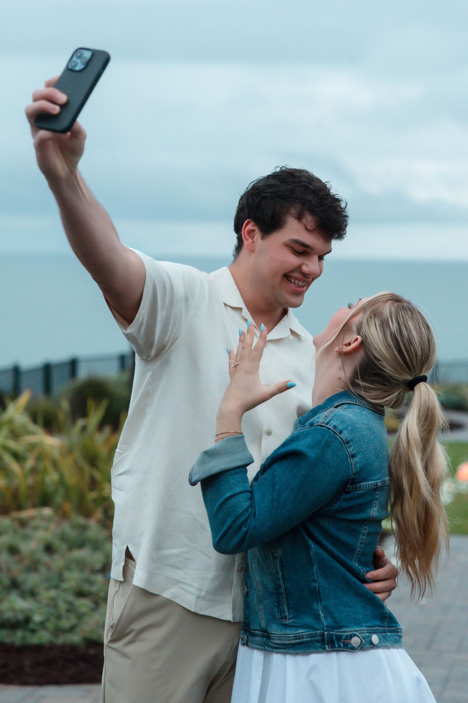 A smiling couple takes a selfie outdoors, with the woman showing off her engagement ring while embracing her partner.