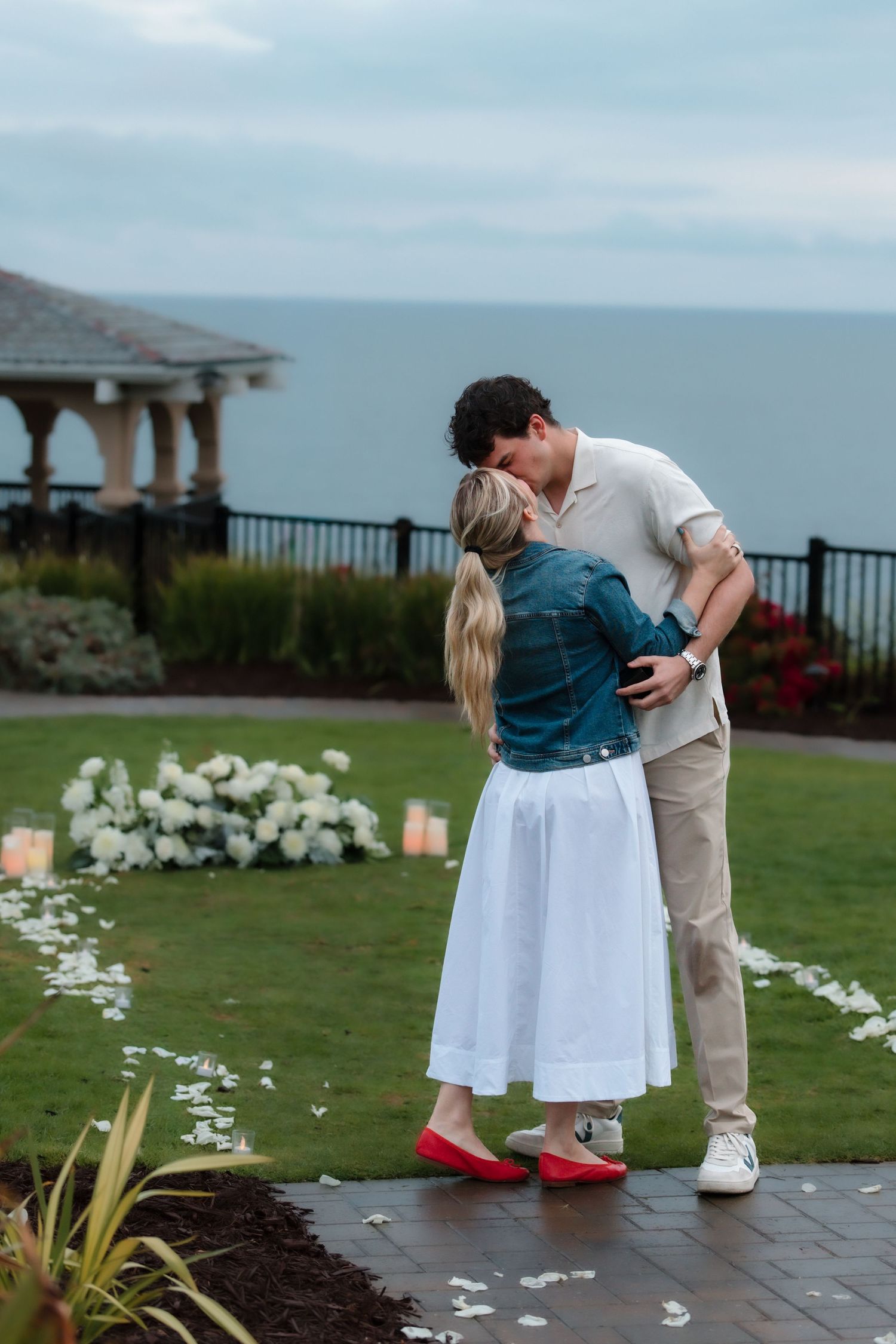 Couple sharing a romantic kiss in a garden with white flowers, candles, and ocean view backdrop.