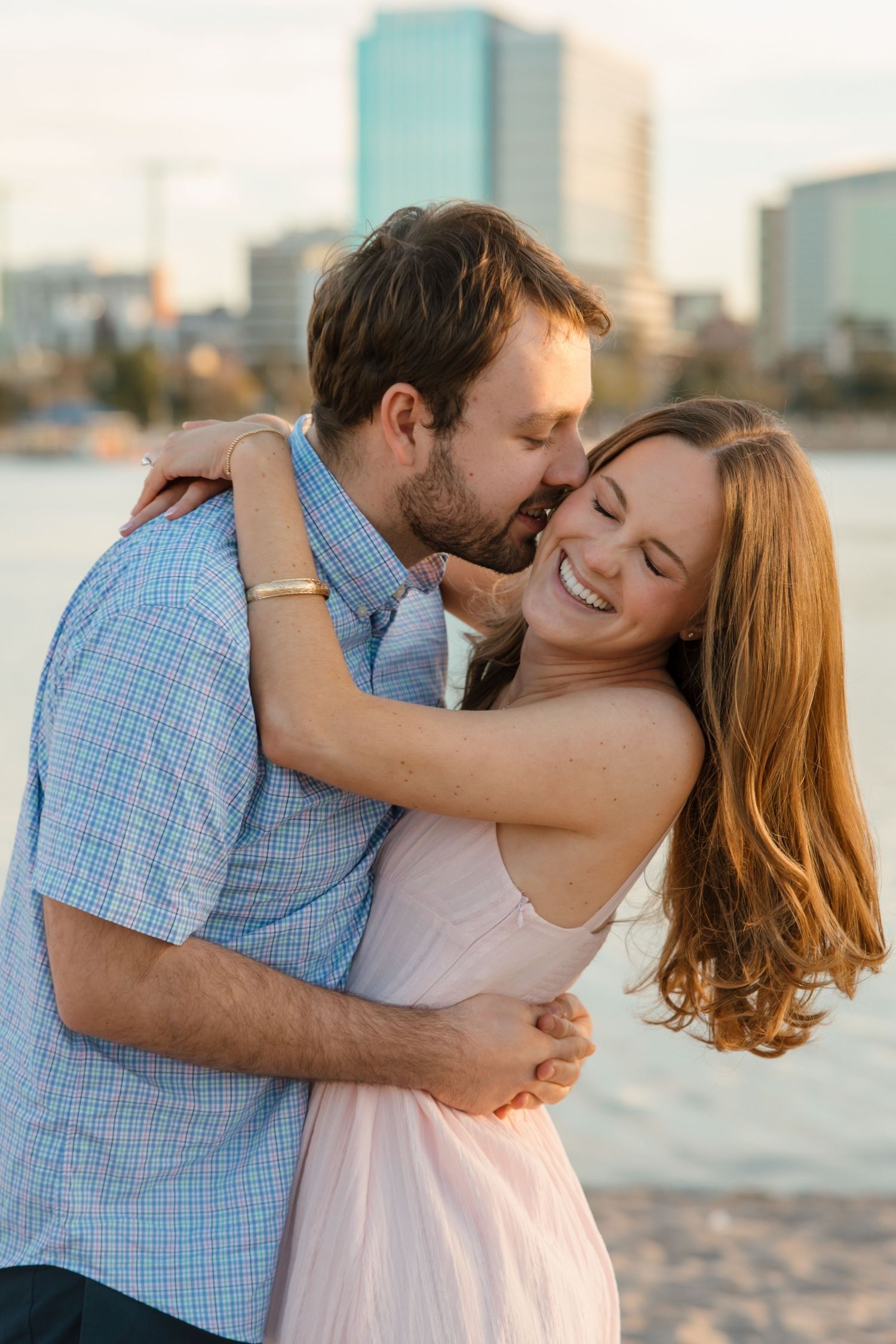 A couple shares a romantic embrace on a waterfront beach with a city skyline backdrop in golden hour light.