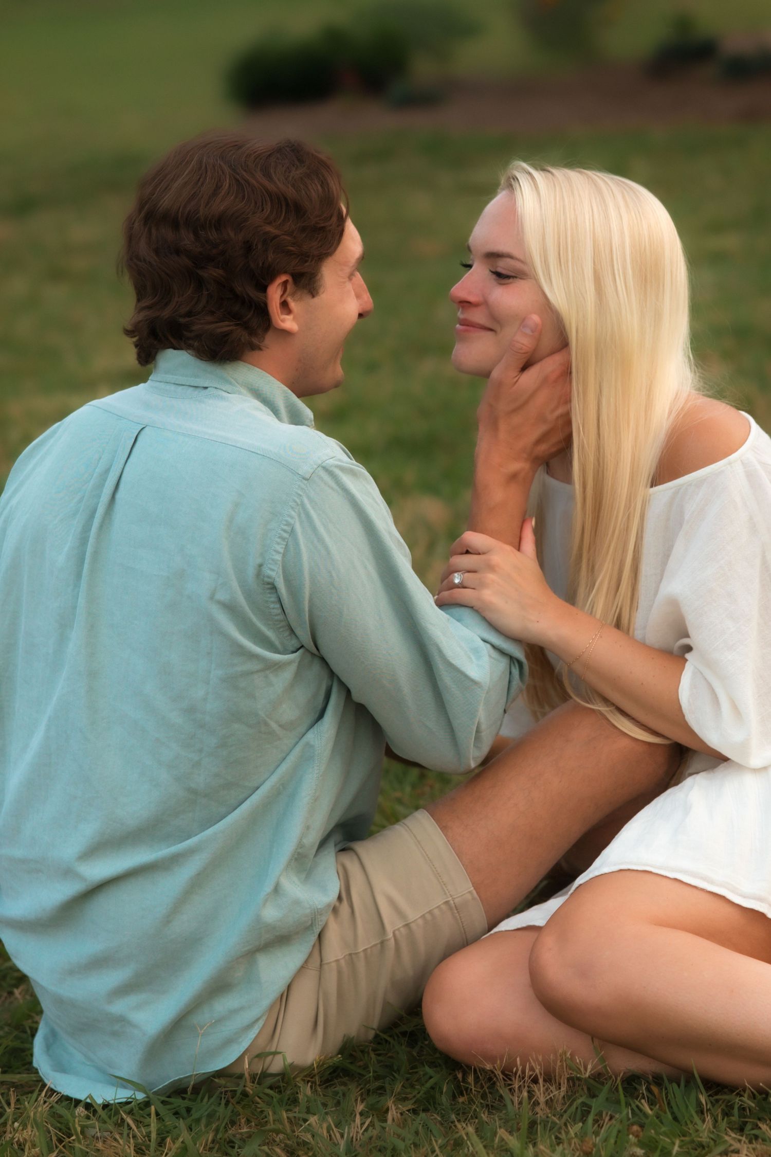 Romantic couple sitting on grass, man gently holding womans face as they gaze into each others eyes.