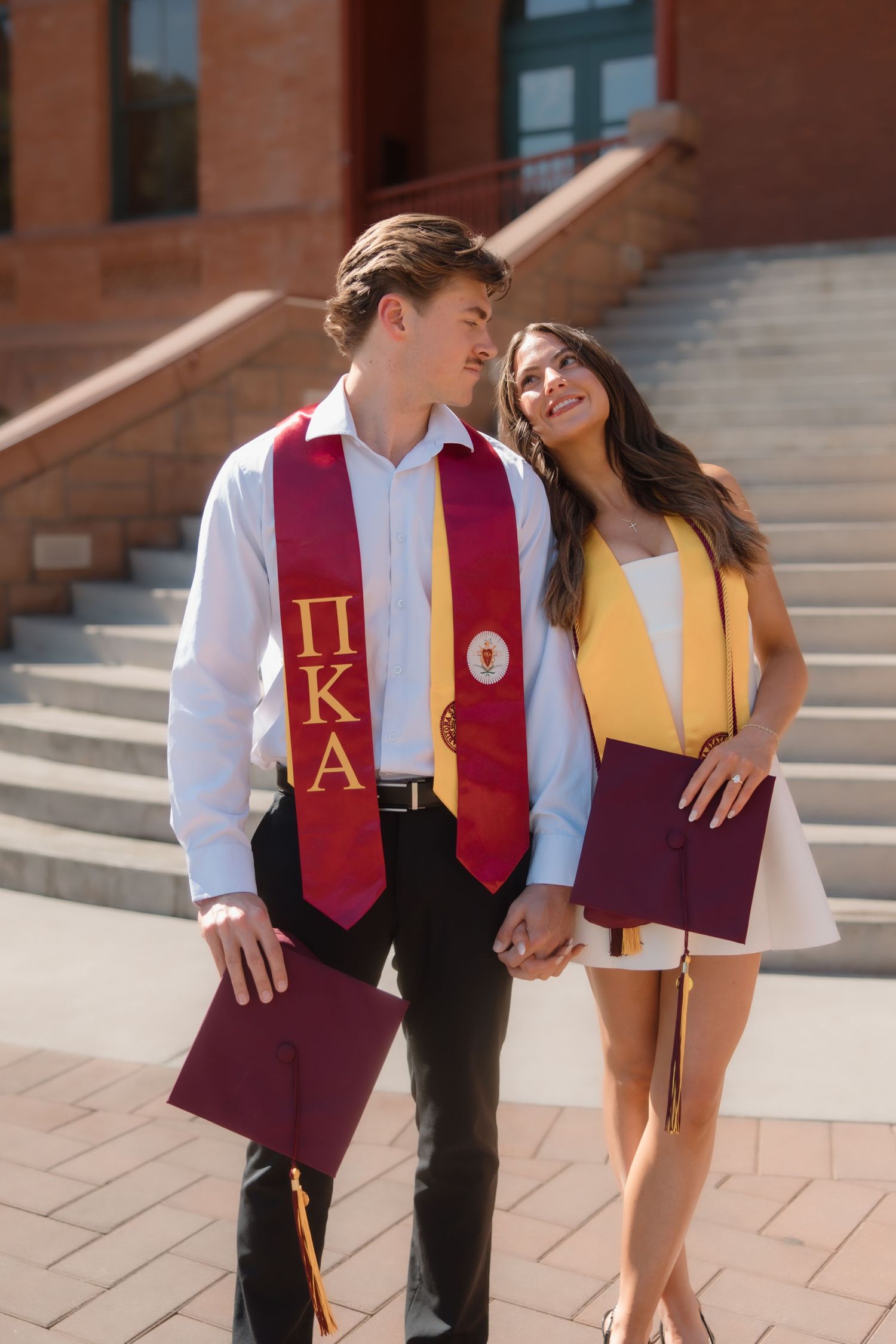Graduating couple holding hands and gazing at each other wearing Pi Kappa Alpha and gold honor stoles.