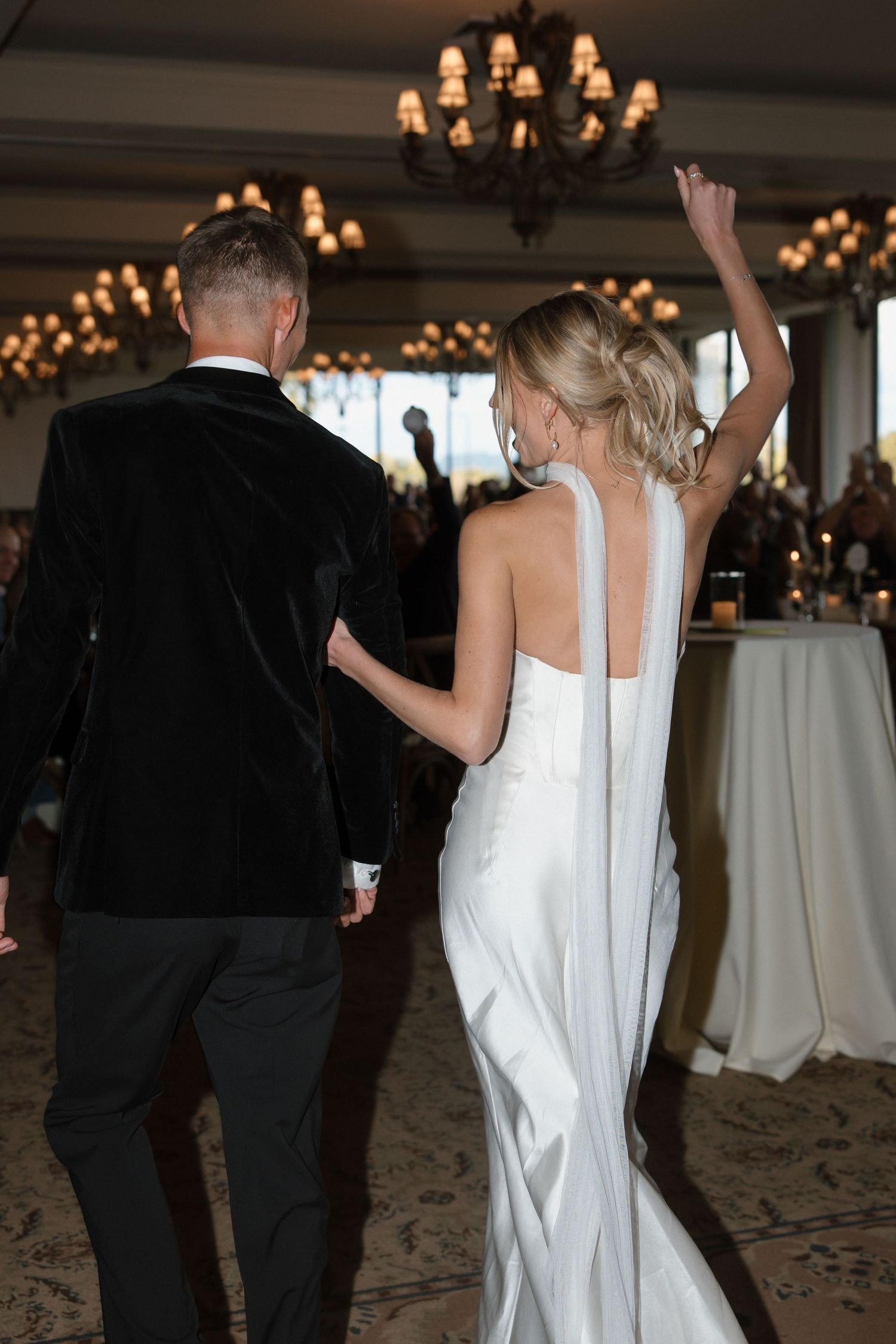 Bride in white halter gown raises arm while walking with groom in black velvet tuxedo at reception.