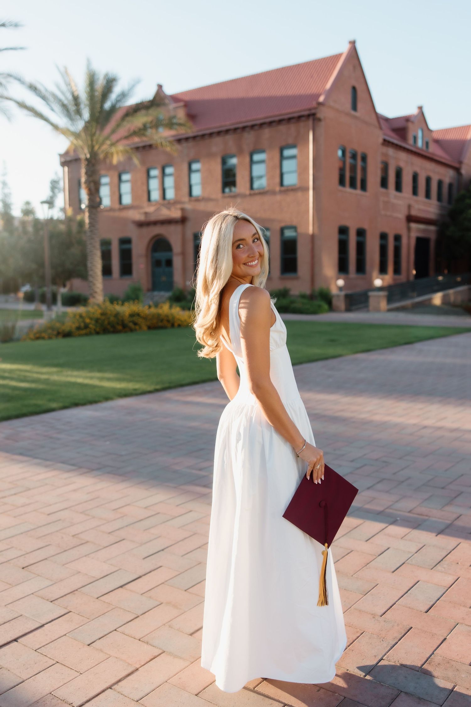 Smiling blonde graduate in white maxi dress holding maroon cap on sunny campus.