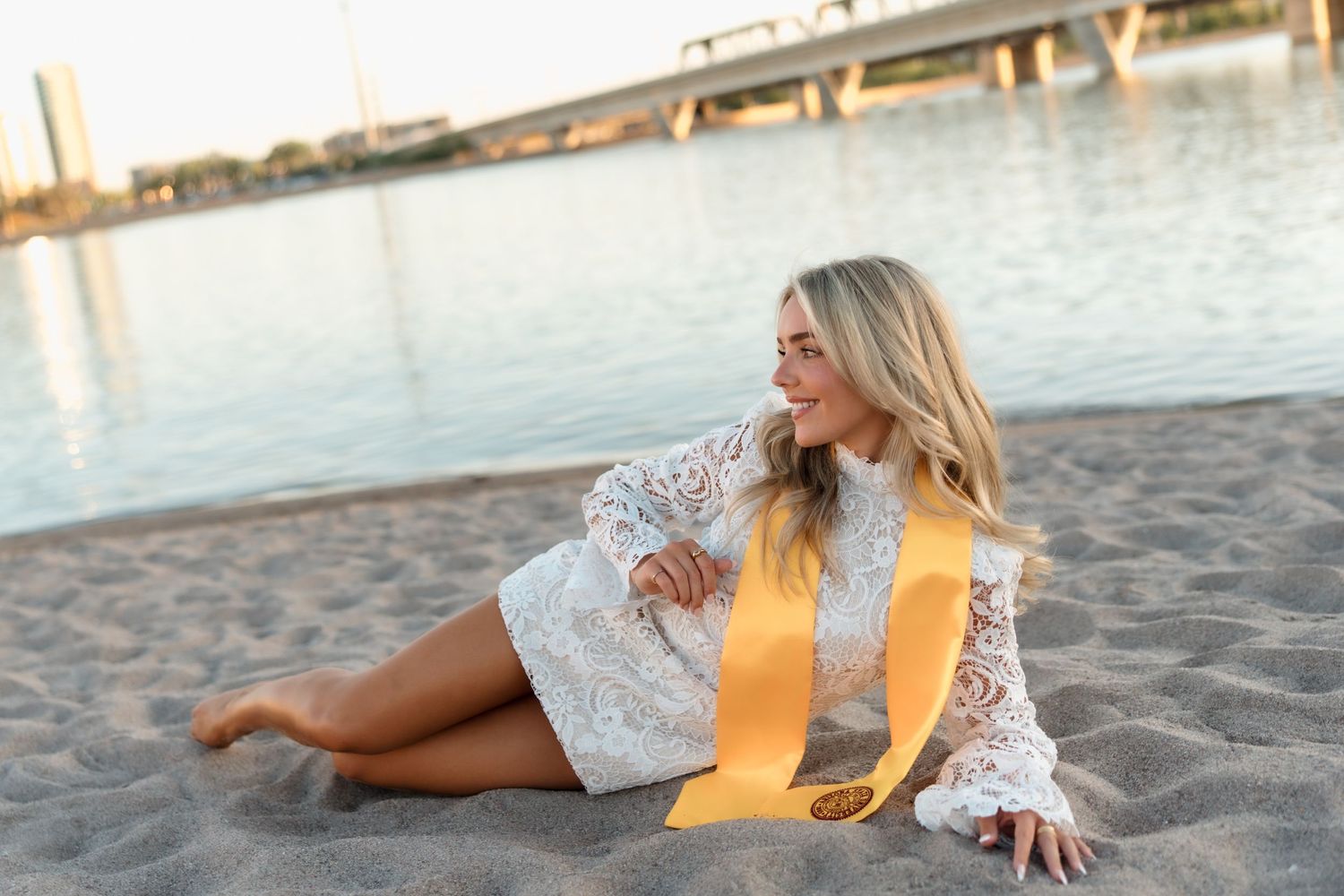 Blonde woman in white lace dress with yellow ribbon sitting on sandy beach by river at sunset.