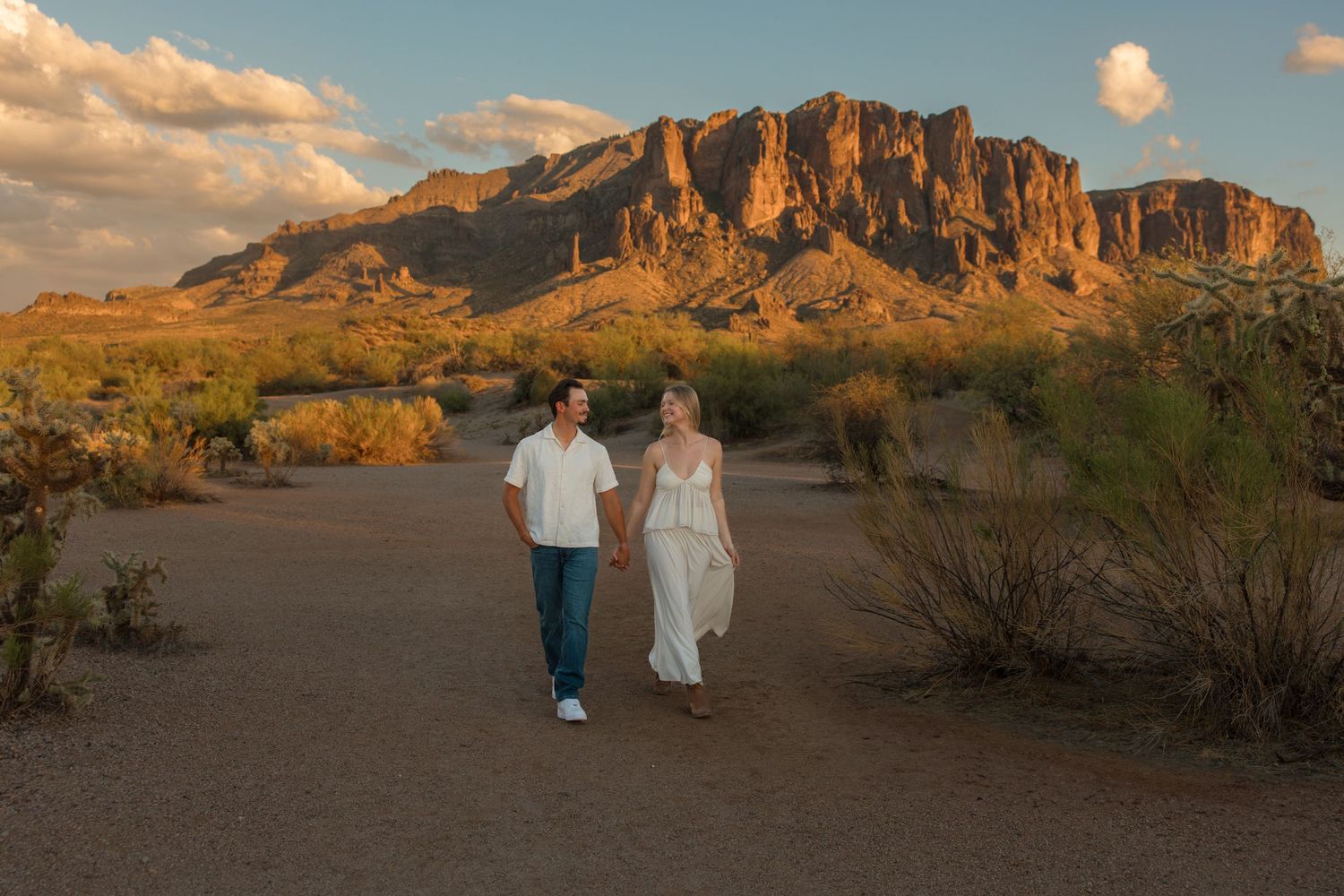 Couple holds hands walking in Superstition Mountains desert landscape at golden hour in Arizona.