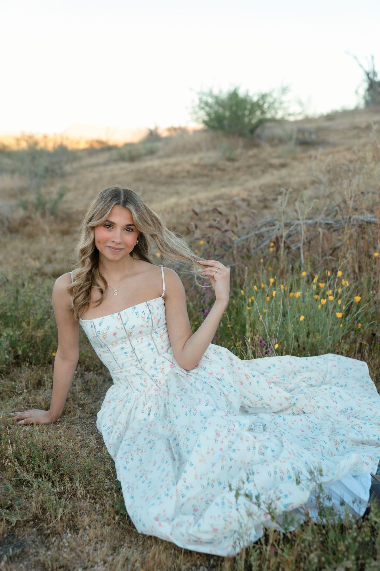 Young woman in floral sundress sitting gracefully in golden meadow at sunset, hair flowing, surrounded by wildflowers.