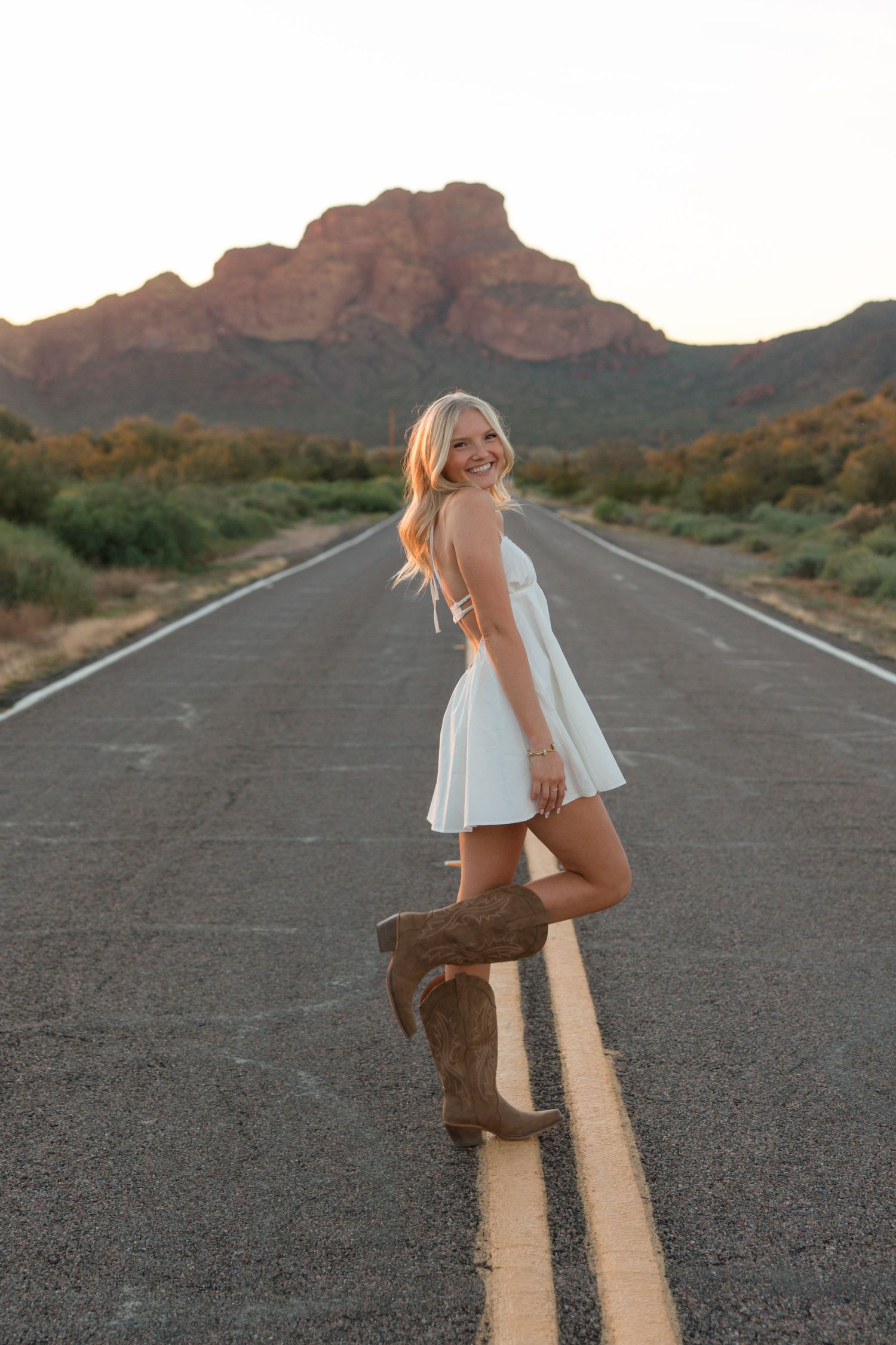 Smiling blonde woman in white dress and cowboy boots posing on desert road with mountain backdrop at sunset.