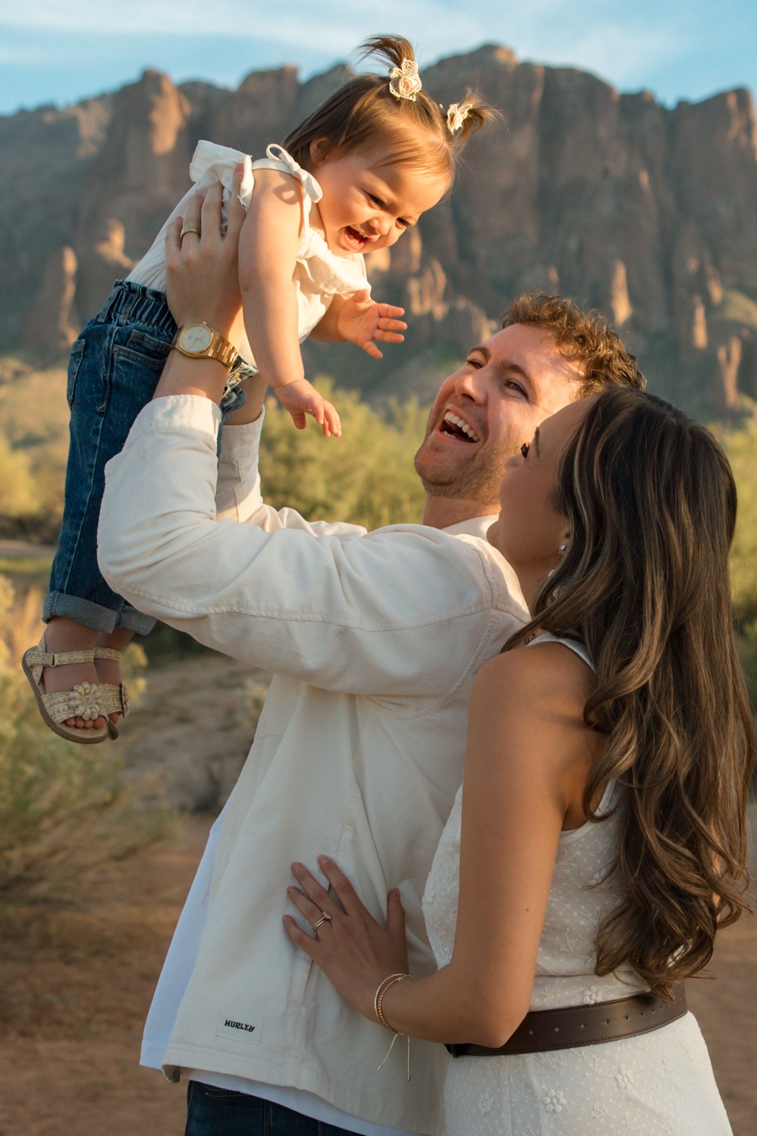 A joyful family with a man lifting a laughing toddler girl while a woman smiles, set against a desert mountain backdrop.