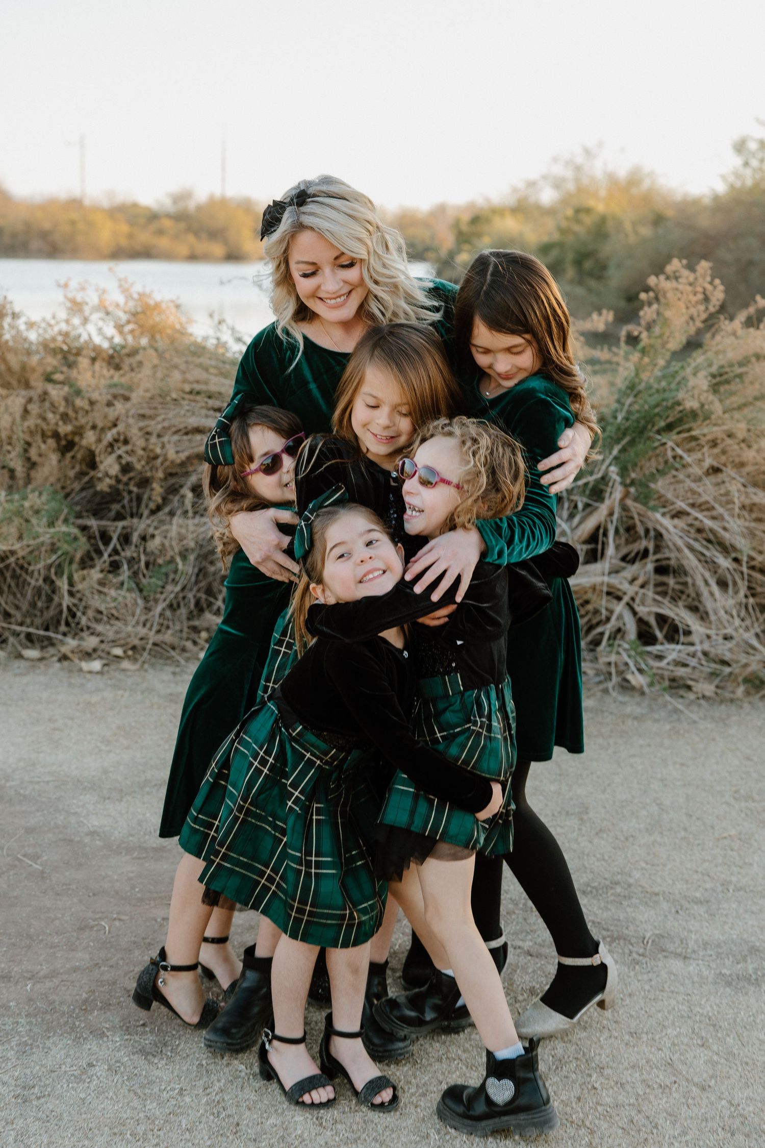 Woman in green velvet dress hugging four girls in matching green plaid dresses outdoors near a river.